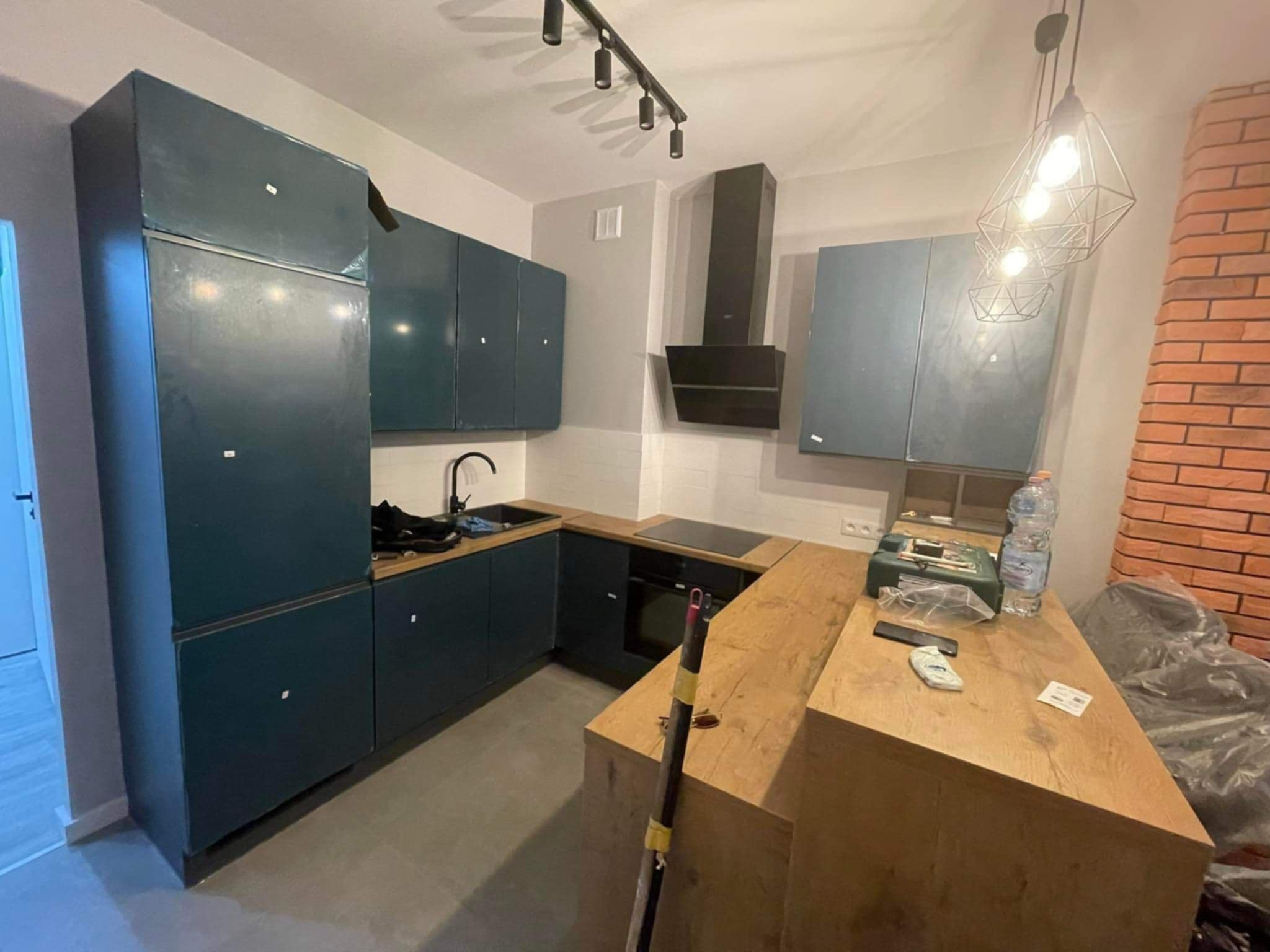 Nowoczesna kuchnia w odcieniach granatu z drewnianym blatem i oświetleniem punktowym. Widoczne akcesoria i narzędzia, sugerujące etap wykończeniowy.