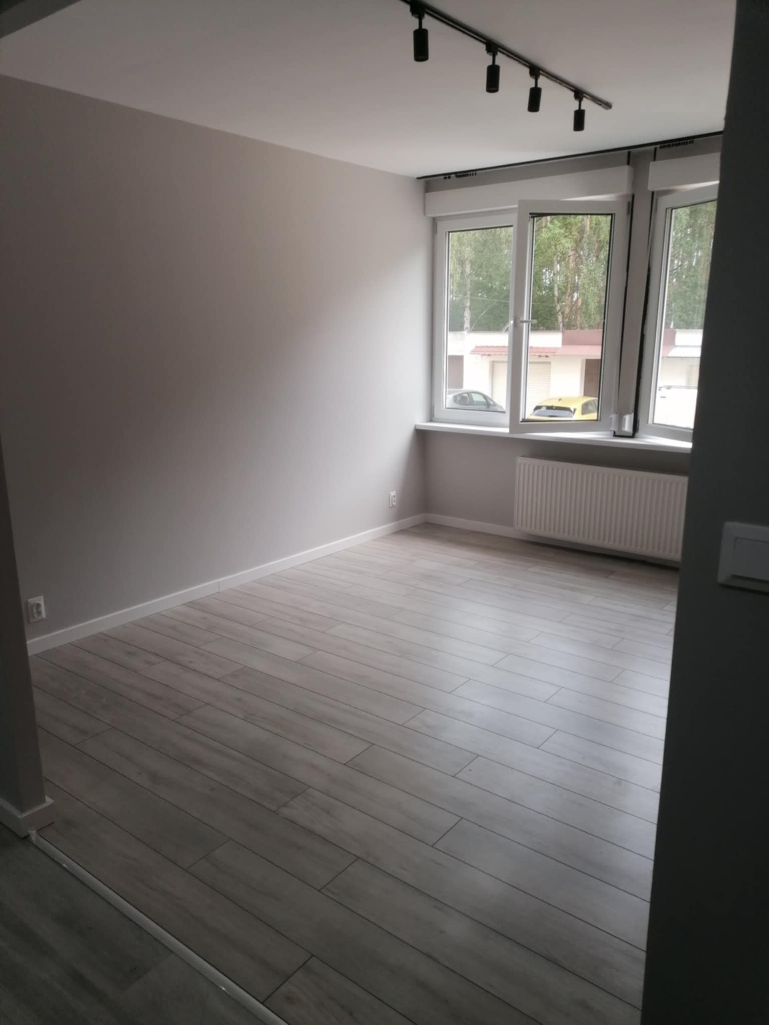 Jasne wnętrze po remoncie: szara podłoga imitująca drewno, ściany w odcieniach szarości, białe okna z widokiem na zieleń. Minimalistyczne oświetlenie sufitowe.