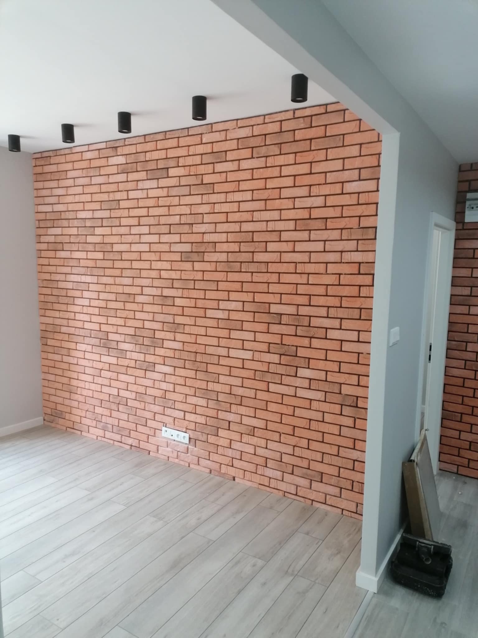 Ściana z dekoracyjnej cegły w odcieniach czerwieni i brązu, oświetlona punktowymi lampami sufitowymi. Jasna podłoga, szara ściana z drzwiami w tle. Widoczne narzędzia.