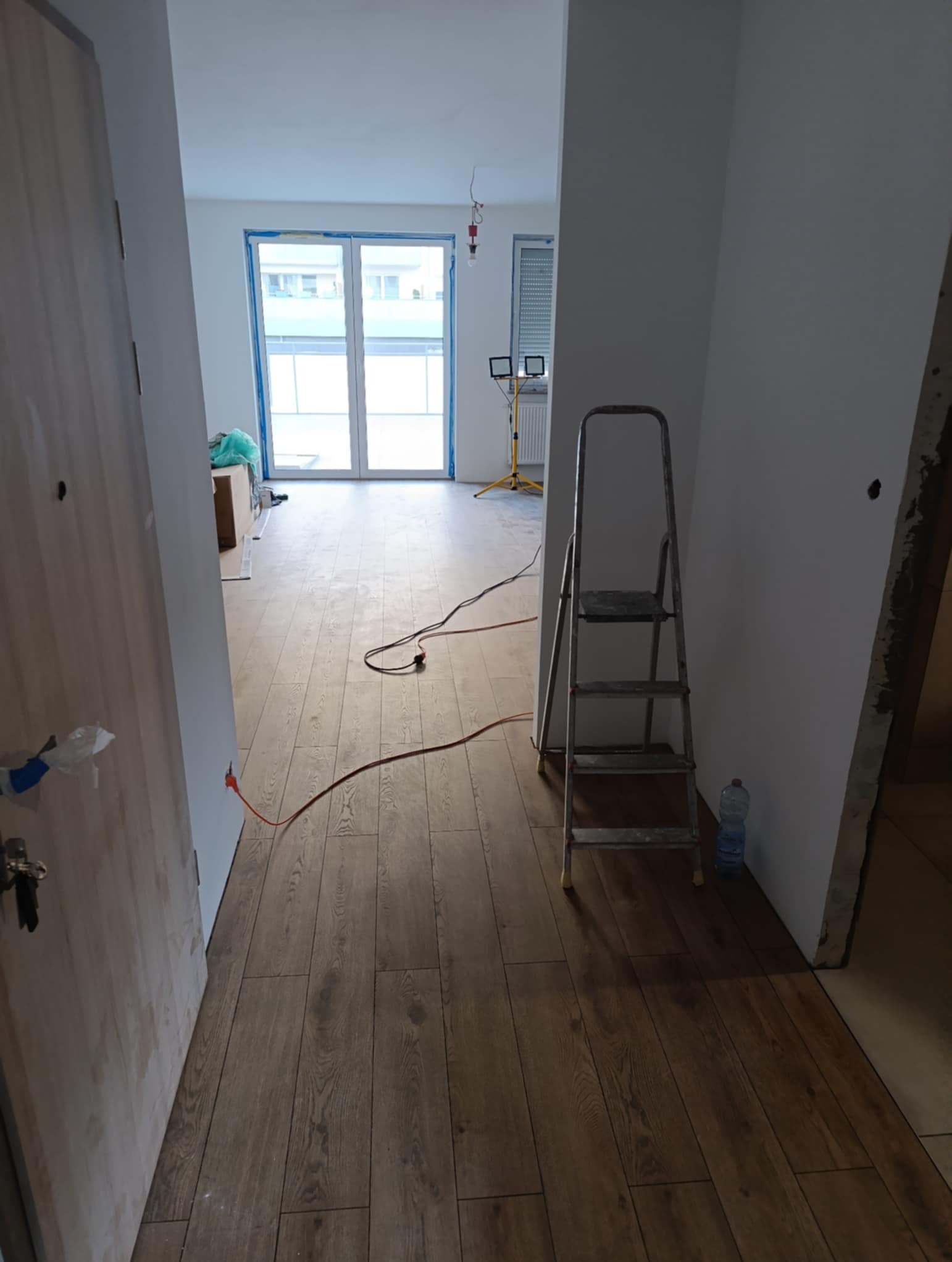 Wnętrze mieszkania w trakcie wykończenia: panele podłogowe ułożone, ściany przygotowane do malowania, widoczna drabina i narzędzia, okno zabezpieczone folią.