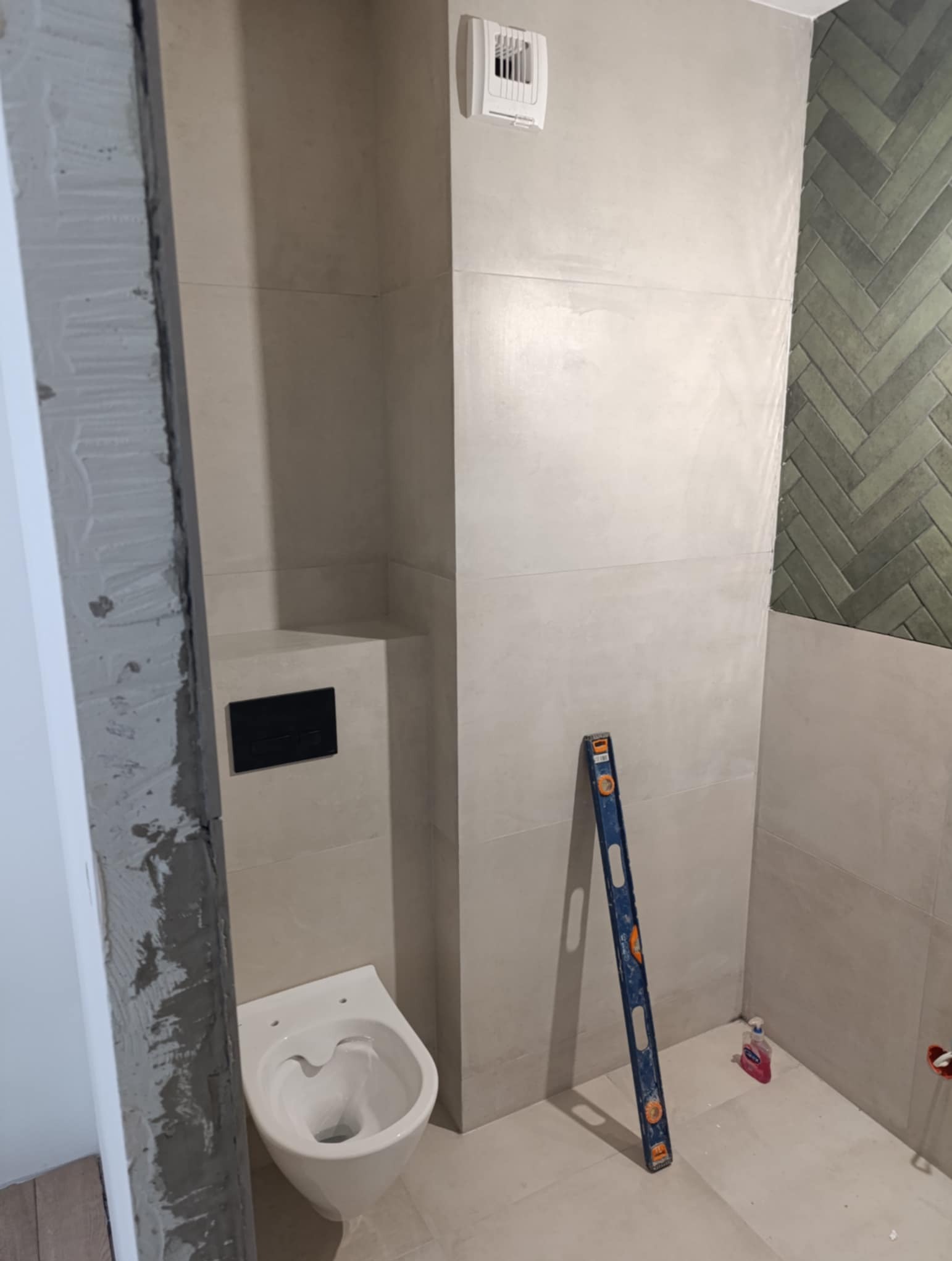 Wykończona łazienka z podwieszaną toaletą, obłożona jasnymi płytkami. Na ścianie zielona mozaika w jodełkę. Widoczna poziomica i fragment ościeżnicy w stanie surowym.