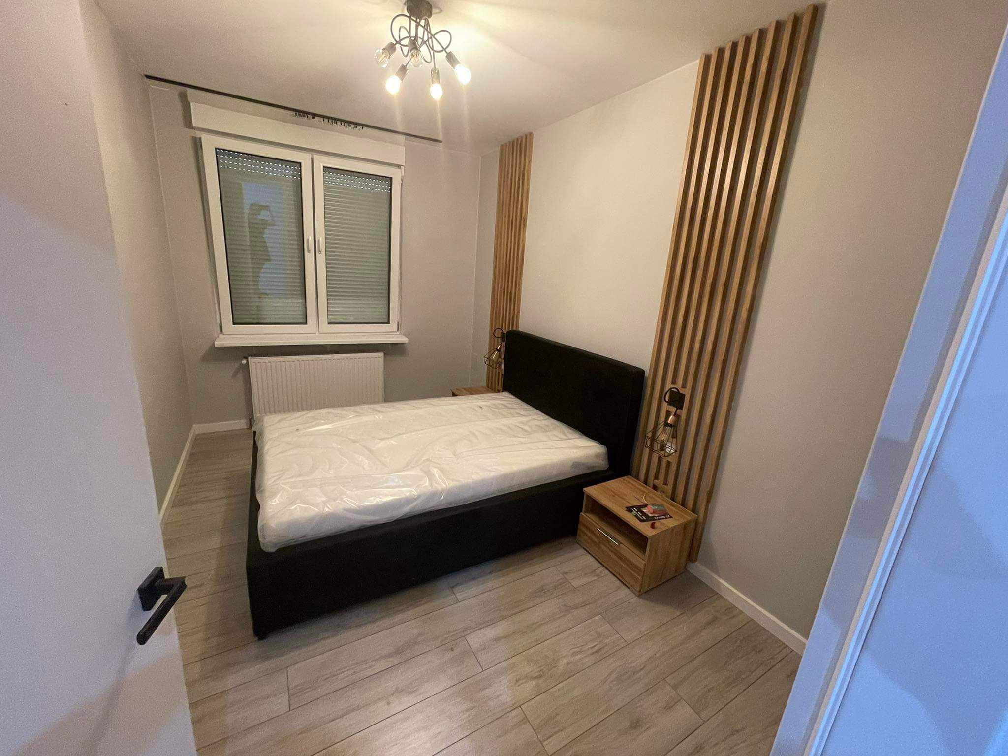 Nowoczesna sypialnia z drewnianymi lamelami na ścianie za łóżkiem. Czarne łóżko z materacem w folii, drewniane podłogi i szafki nocne. Minimalistyczny wystrój.