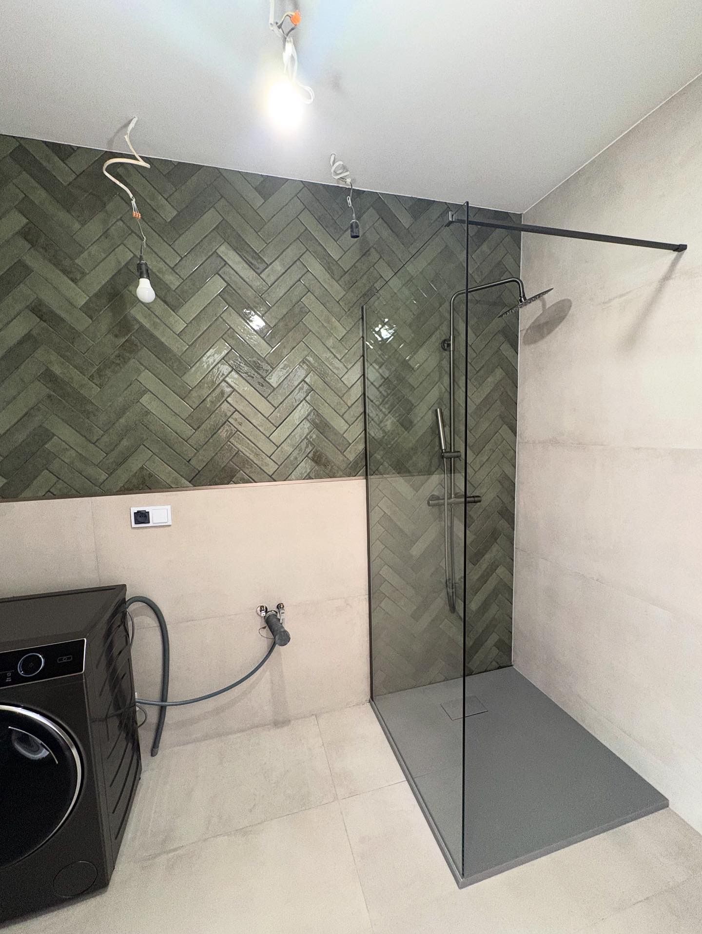 Nowoczesna łazienka z kabiną prysznicową typu walk-in, wykończona zieloną glazurą ułożoną w jodełkę i szarą posadzką. Widoczna pralka i niedokończone oświetlenie.