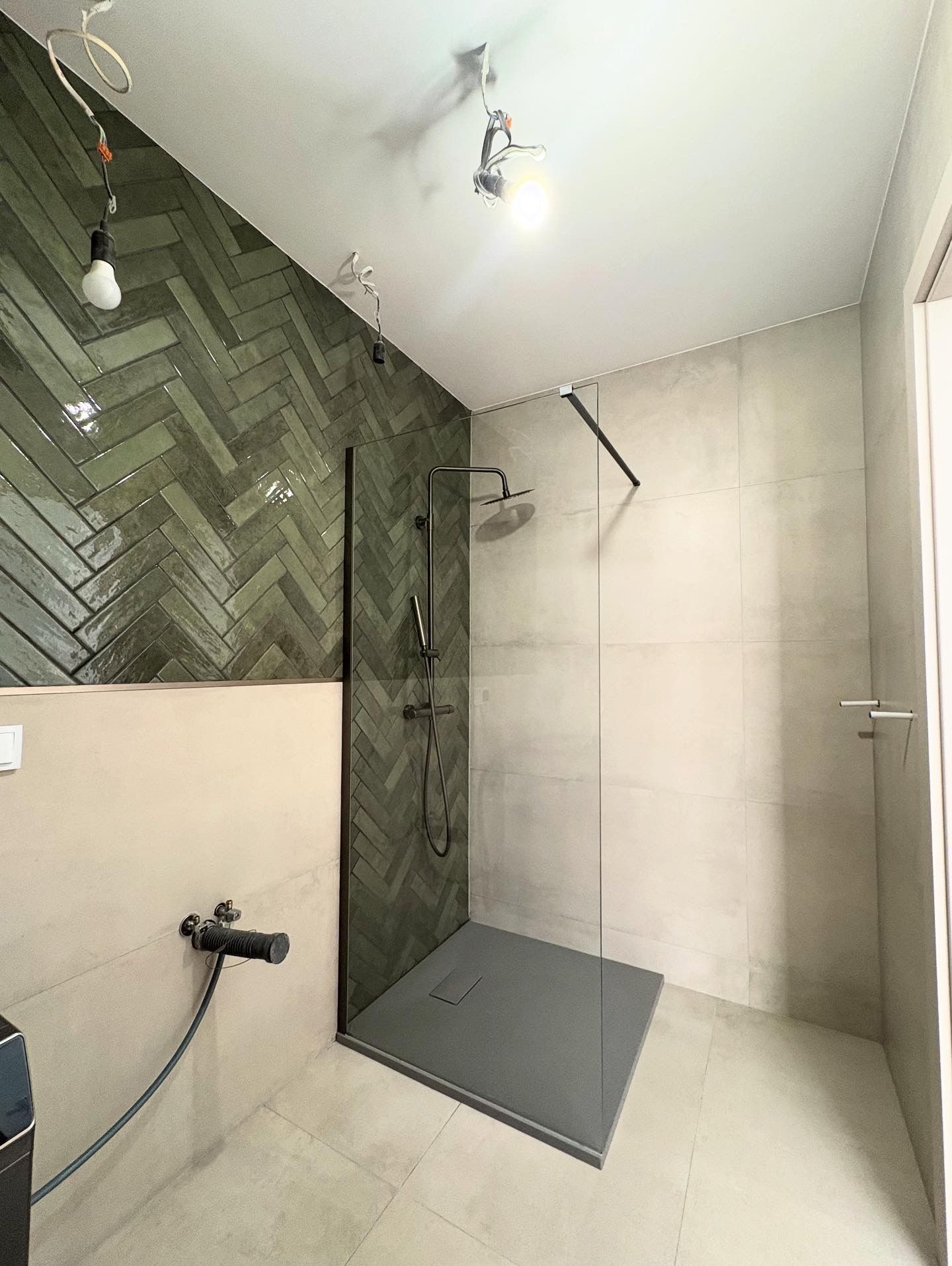 Nowoczesna łazienka z kabiną prysznicową walk-in, wykończona zieloną, ułożoną w jodełkę glazurą i beżowymi płytkami. Widoczne niedokończone oświetlenie sufitowe.