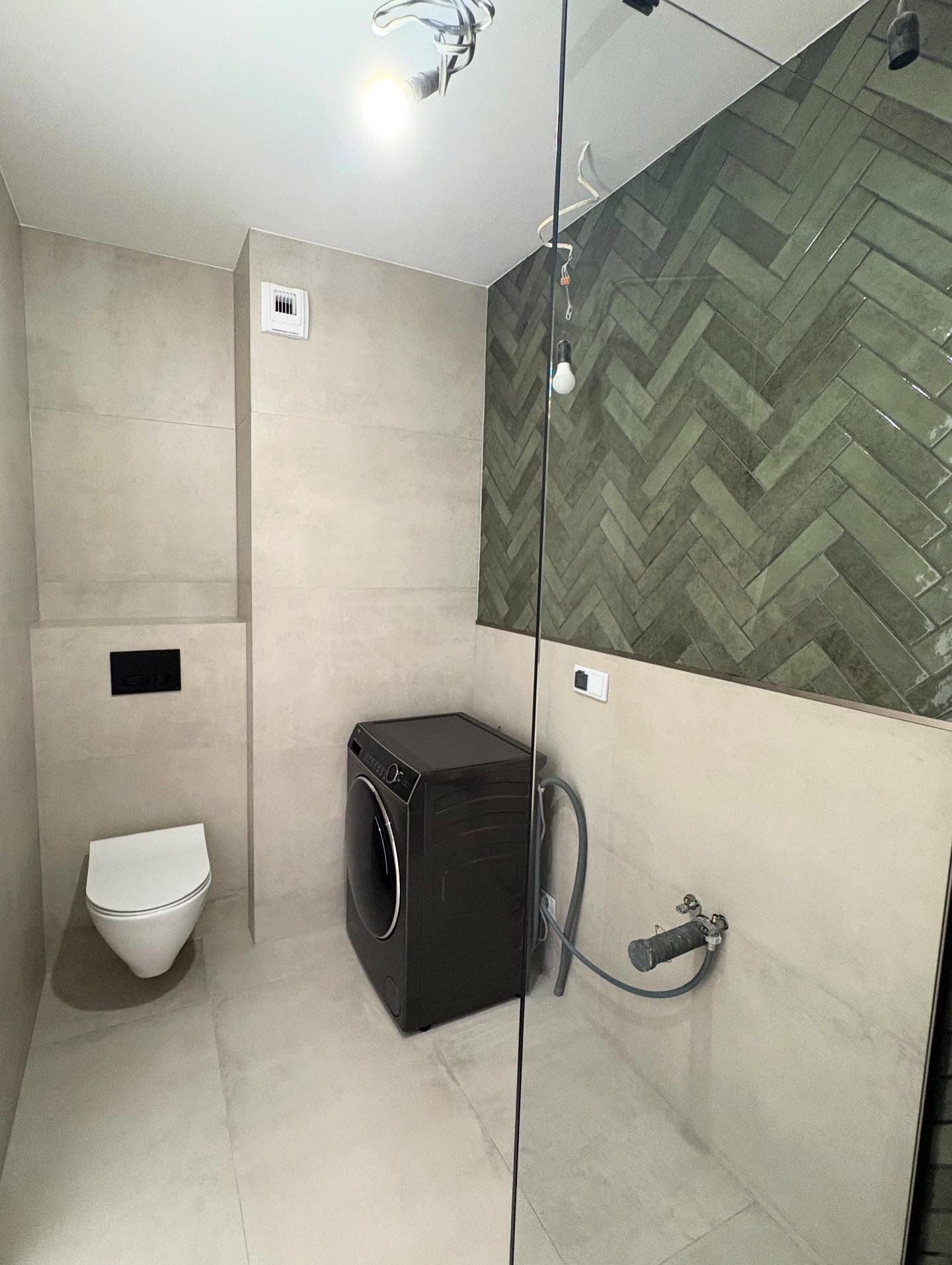 Nowoczesna łazienka z toaletą, pralką i zielonymi płytkami ułożonymi w jodełkę na ścianie. Widoczne szklane elementy kabiny prysznicowej i oświetlenie sufitowe.