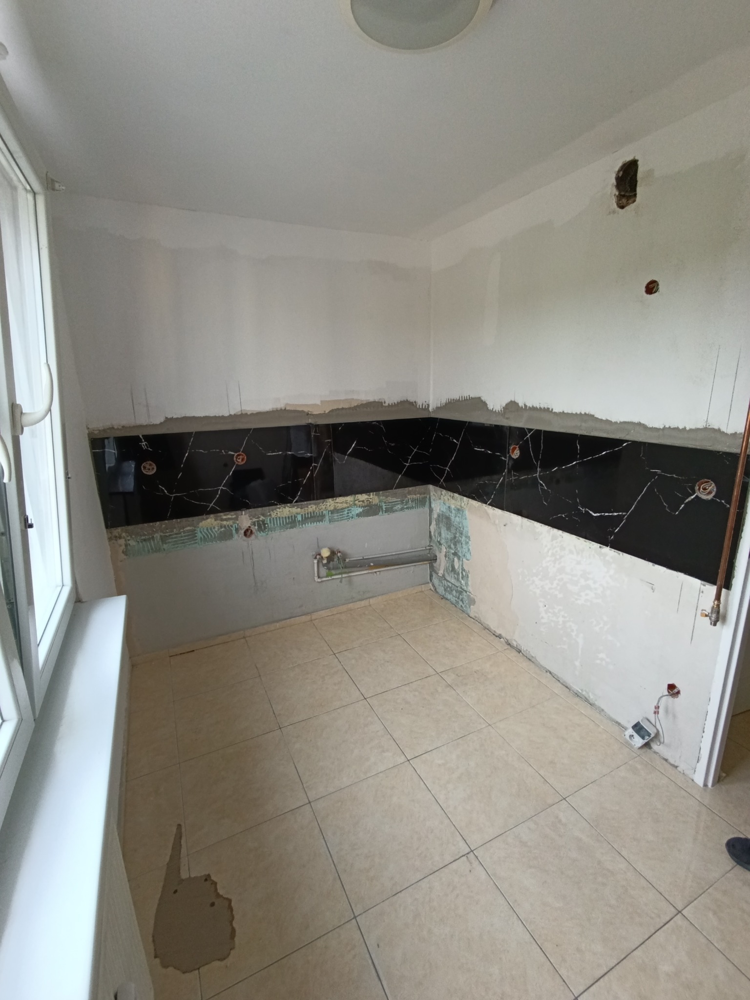 Częściowo wykończona kuchnia: czarne płytki z białym wzorem marmuru na ścianie, beżowe płytki na podłodze, ściany przygotowane do dalszych prac. Widoczne instalacje.