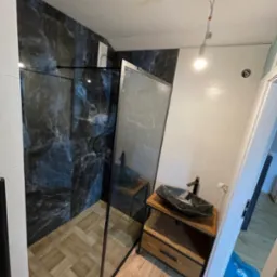 Wykończona łazienka z kabiną typu walk-in, marmurowymi płytkami i drewnianą szafką z umywalką w stylu loft. Widoczny fragment podłogi z drewnopodobnych płytek.