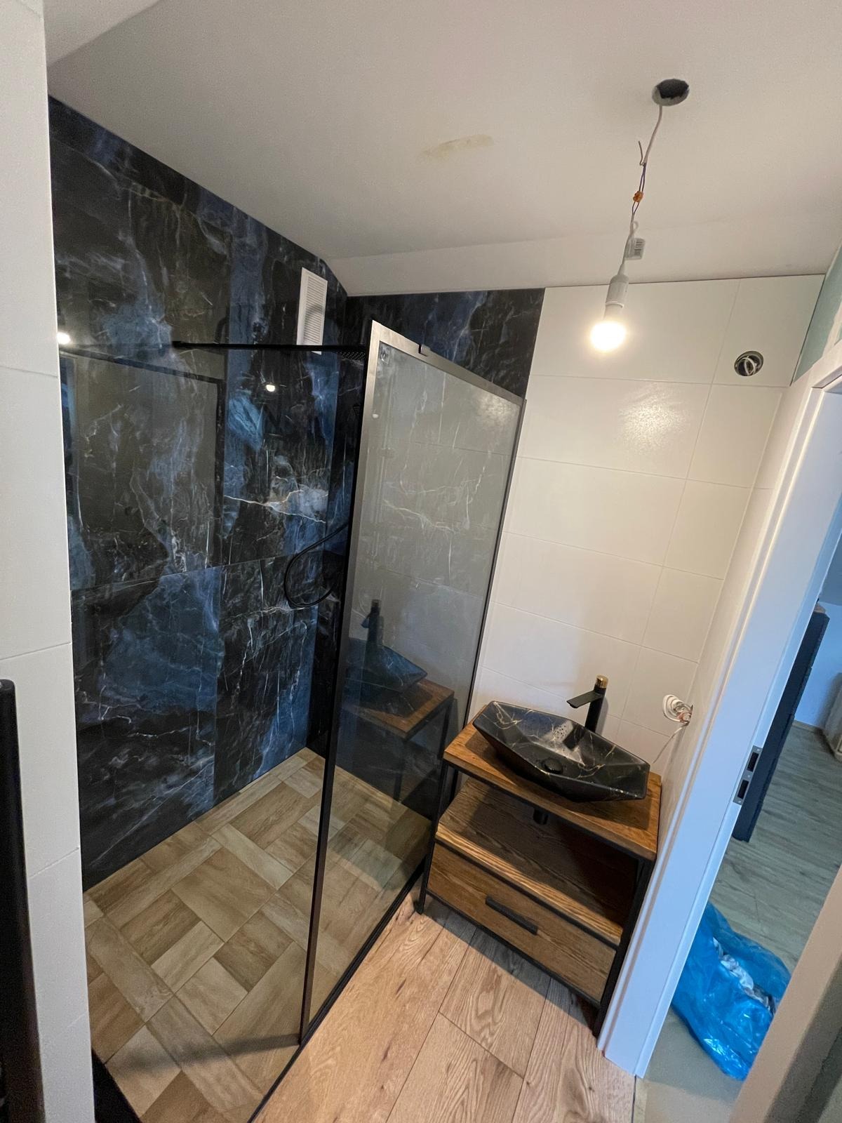 Wykończona łazienka z kabiną typu walk-in, marmurowymi płytkami i drewnianą szafką z umywalką w stylu loft. Widoczny fragment podłogi z drewnopodobnych płytek.