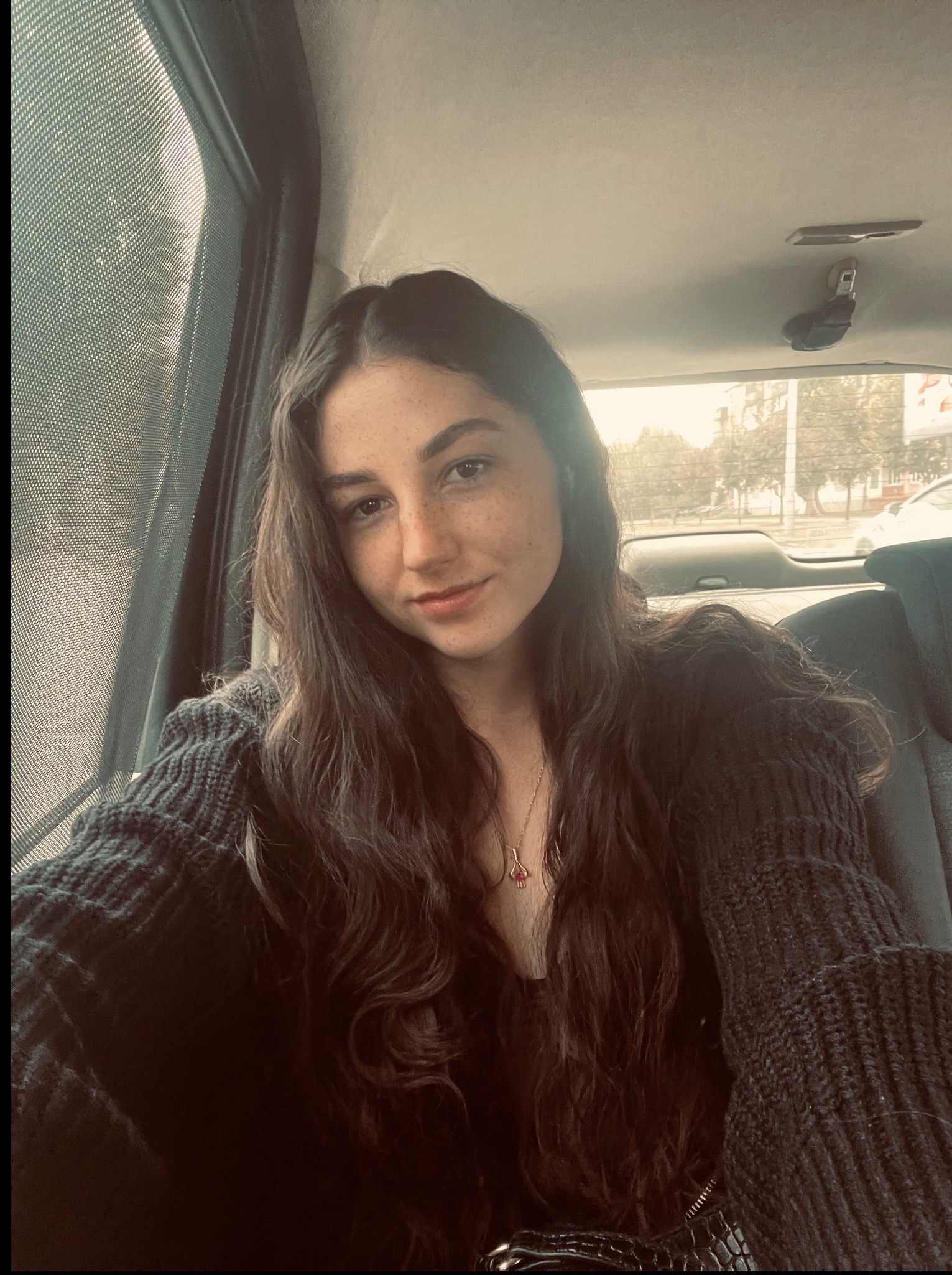 Portret kobiety z piegami, siedzącej w samochodzie, z widokiem na miasto za oknem. Delikatne światło podkreśla jej naturalny wygląd i swobodną pozę.