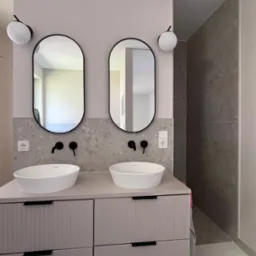 Nowoczesna łazienka z dwoma umywalkami nablatowymi i owalnymi lustrami w czarnych ramach, oświetlona kulistymi lampami. Szara mozaika na ścianie i minimalistyczne baterie.