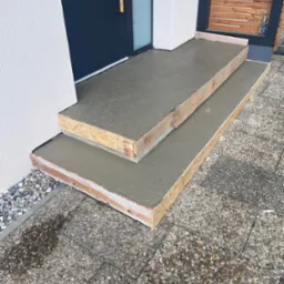 Betonowe schody wejściowe w drewnianym szalunku, świeżo wylane, z widoczną fakturą betonu i otoczeniem z kostki brukowej. Widoczny fragment drzwi wejściowych.