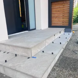 Montaż płytek na schodach wejściowych do domu. Użyto poziomicy i krzyżyków dystansowych. Widoczna drewniana konstrukcja obok schodów i żwirowe podłoże.