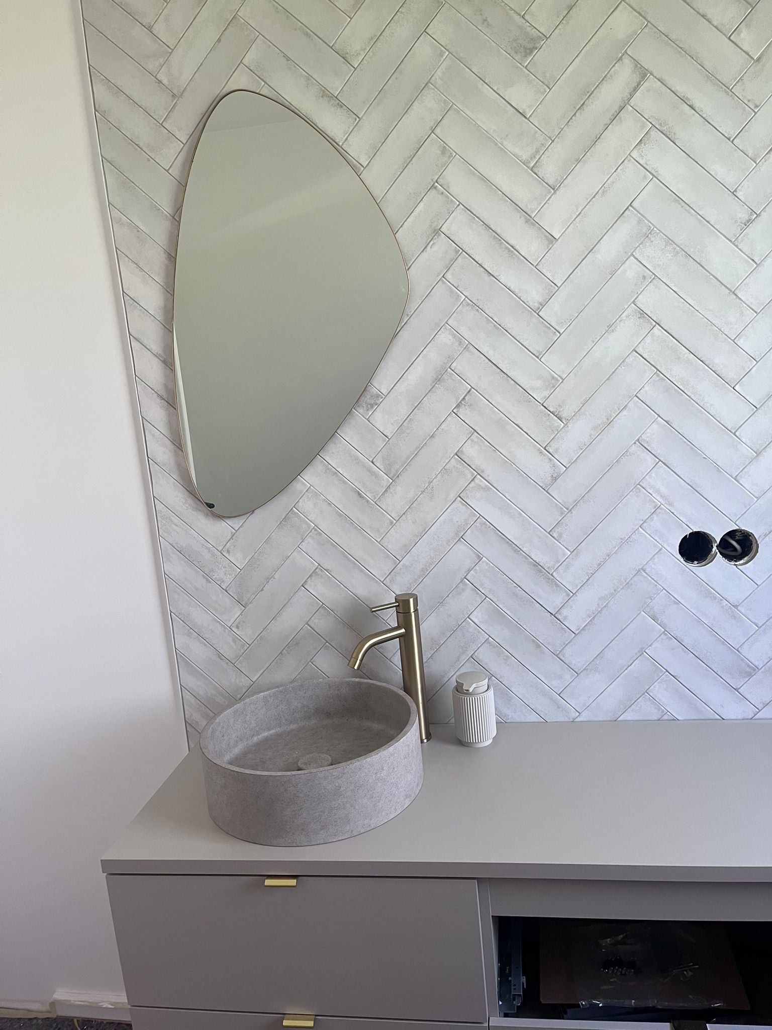Nowoczesna łazienka z umywalką w kształcie misy i lustrem o nieregularnym kształcie na ścianie z białej cegły ułożonej w jodełkę. Złota bateria i szara szafka pod umywalką.
