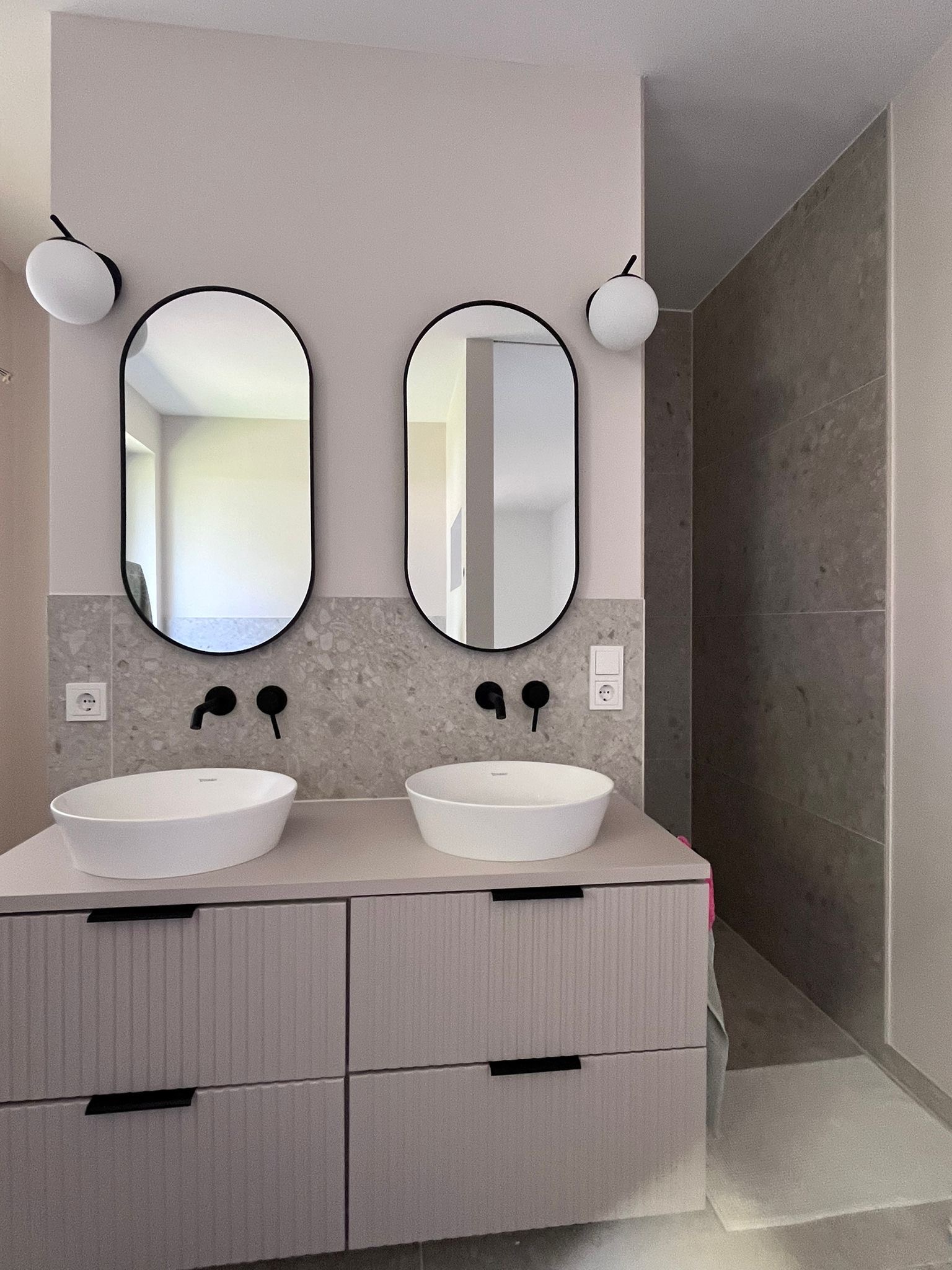 Nowoczesna łazienka z dwoma umywalkami nablatowymi i owalnymi lustrami w czarnych ramach, oświetlona kulistymi lampami. Szara mozaika na ścianie i minimalistyczne baterie.