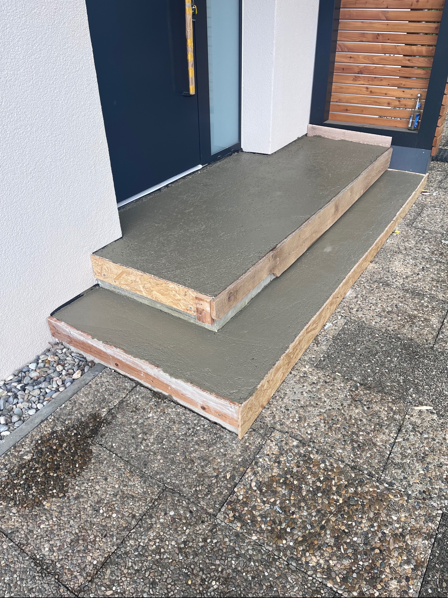 Betonowe schody wejściowe w drewnianym szalunku, świeżo wylane, z widoczną fakturą betonu i otoczeniem z kostki brukowej. Widoczny fragment drzwi wejściowych.