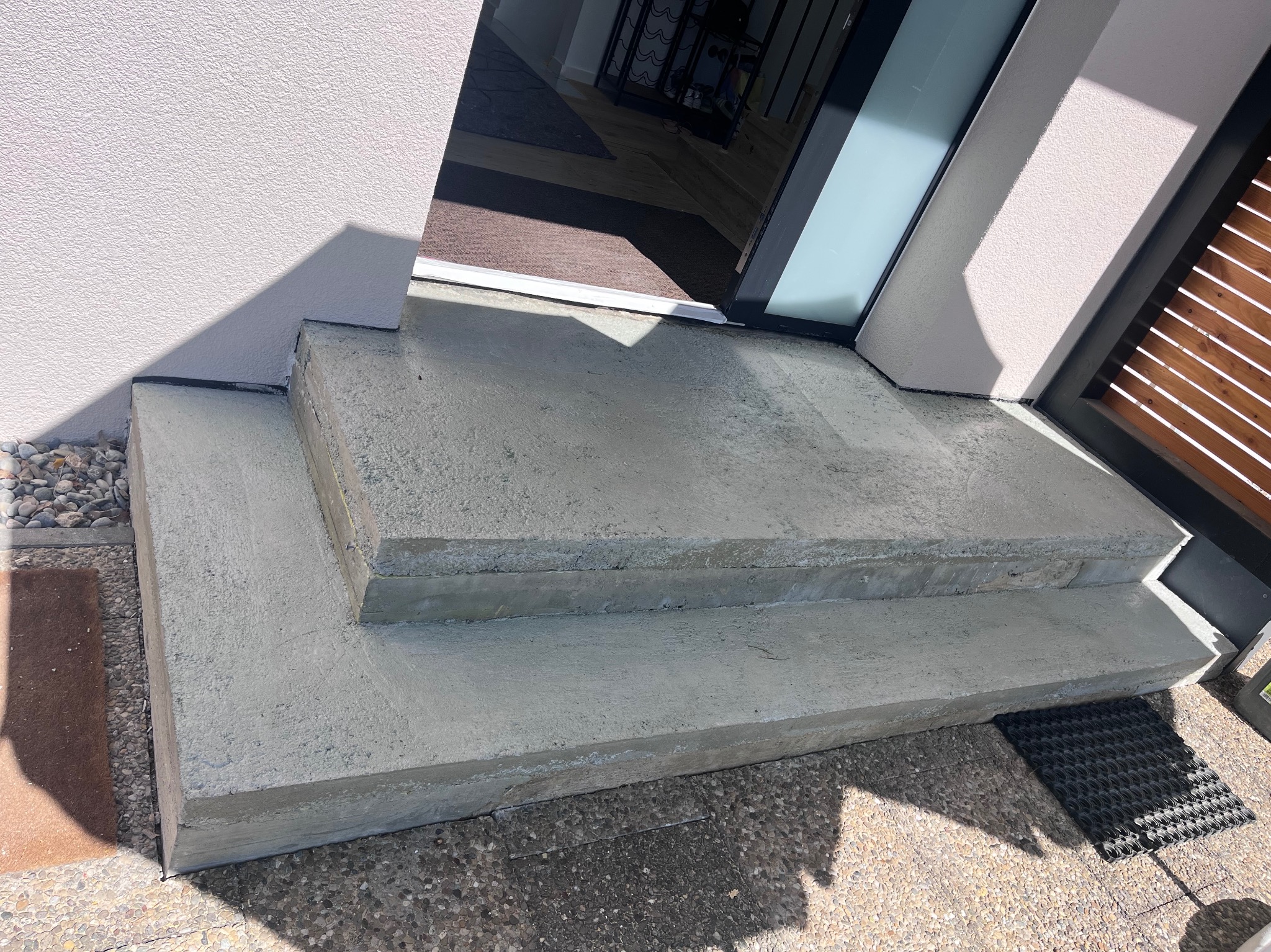 Betonowe schody wejściowe do budynku z teksturą, widoczne z bliska, z fragmentem drzwi i elewacji w tle, oświetlone naturalnym światłem.