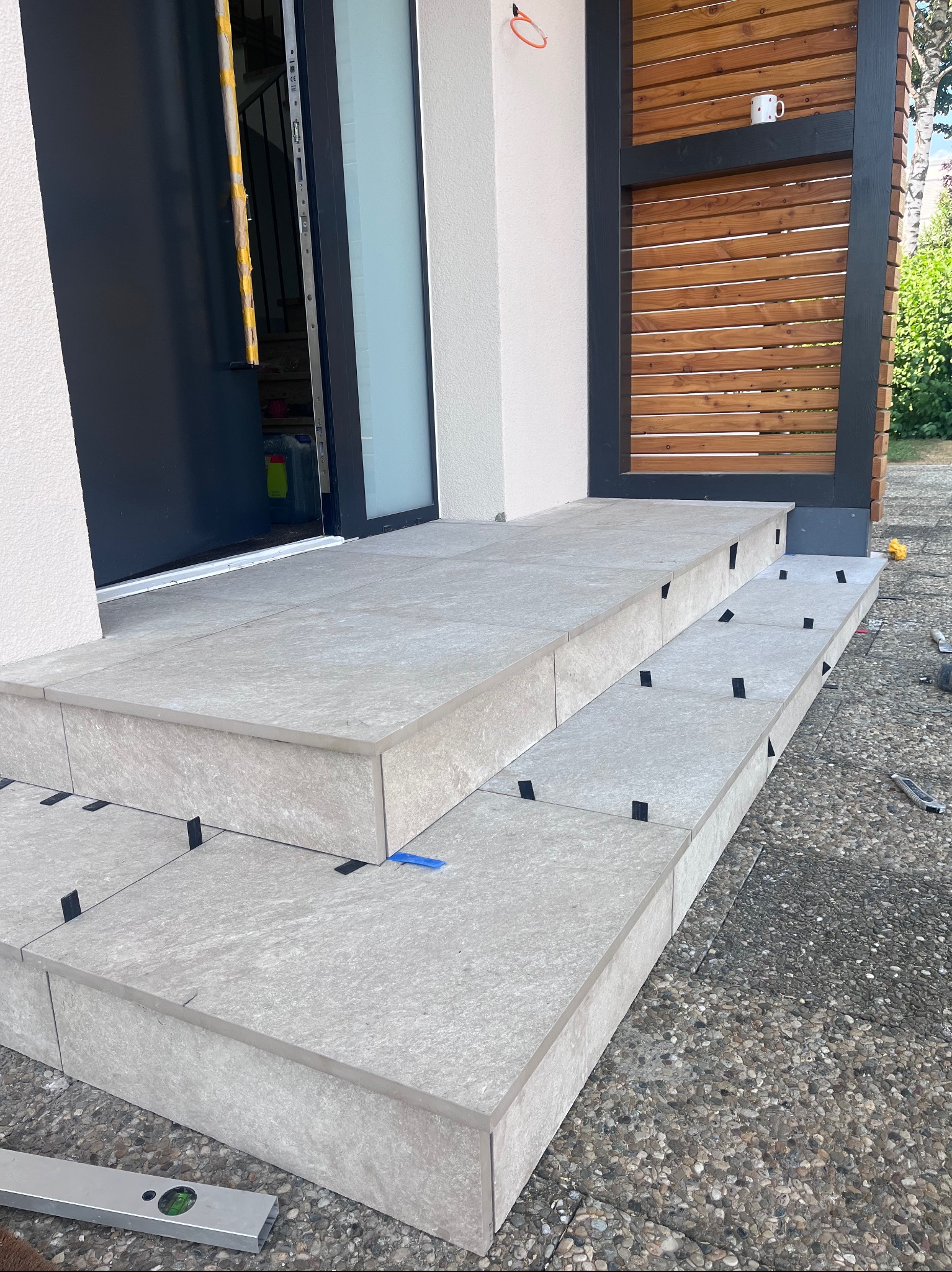 Montaż płytek na schodach wejściowych do domu. Użyto poziomicy i krzyżyków dystansowych. Widoczna drewniana konstrukcja obok schodów i żwirowe podłoże.