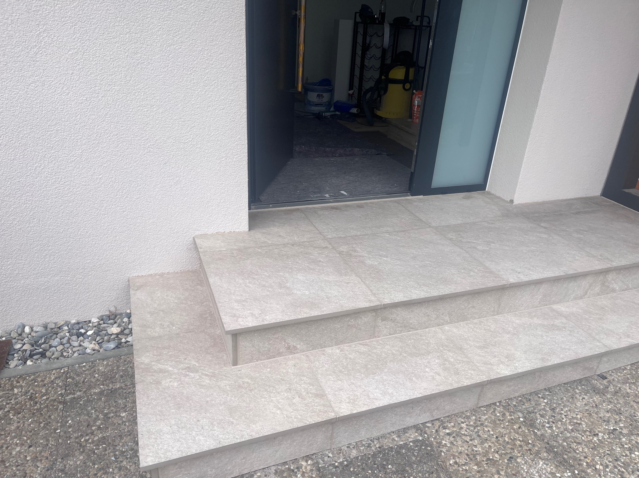 Wykończone schody wejściowe z jasnych, kwadratowych płytek, prowadzące do otwartych drzwi. Widoczne małe kamienie dekoracyjne obok schodów.