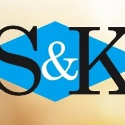 Logo 'S&K' w czcionce szeryfowej, czarne litery na błękitnym rombie, na rozmytym, ciepłym tle. Minimalistyczny design logo.
