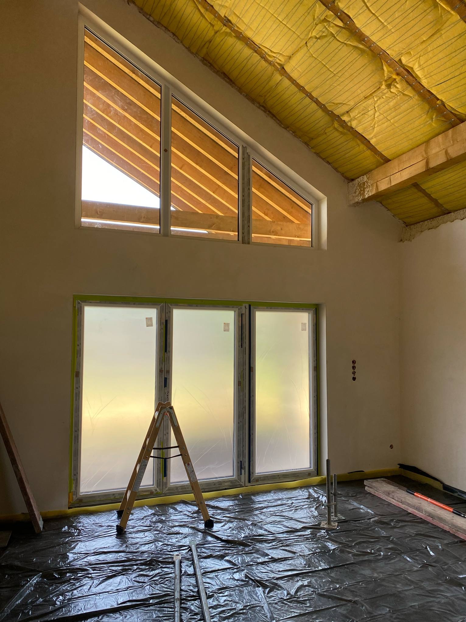 Wnętrze w trakcie prac: okna zabezpieczone folią, podłoga przykryta czarną folią ochronną, widoczne rusztowania i izolacja stropu z żółtej wełny mineralnej.