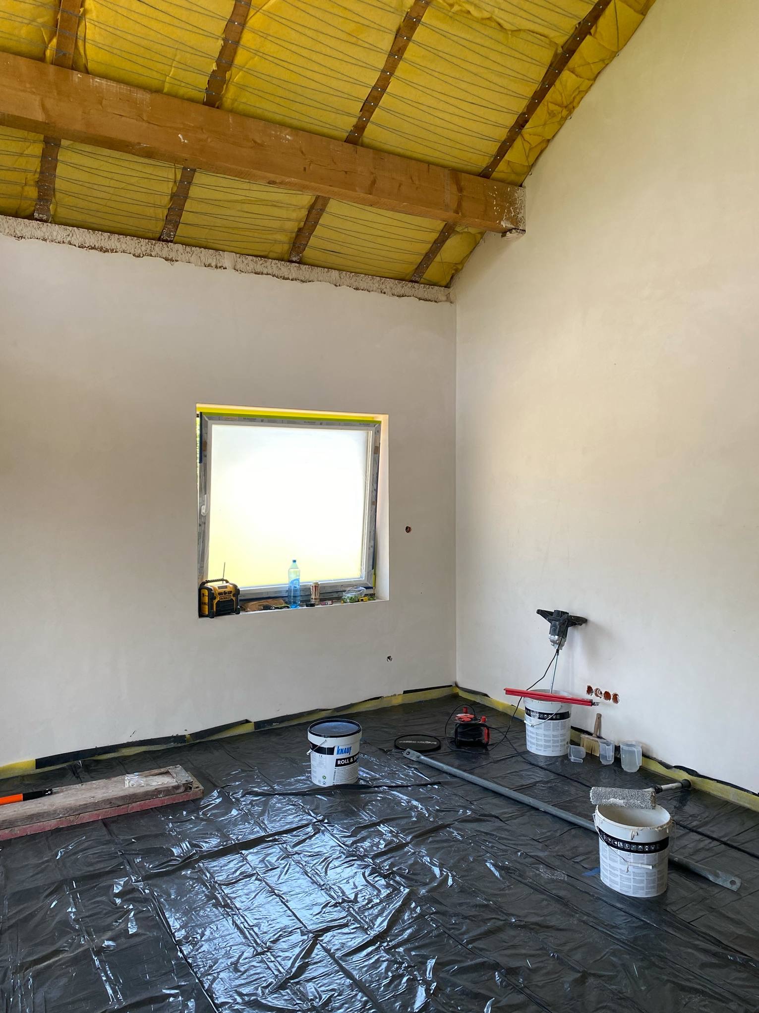 Pokój w trakcie prac wykończeniowych: świeżo nałożona gładź na ścianach, okno z żółtą folią, podłoga zabezpieczona folią, widoczne narzędzia i puszki z gładzią.