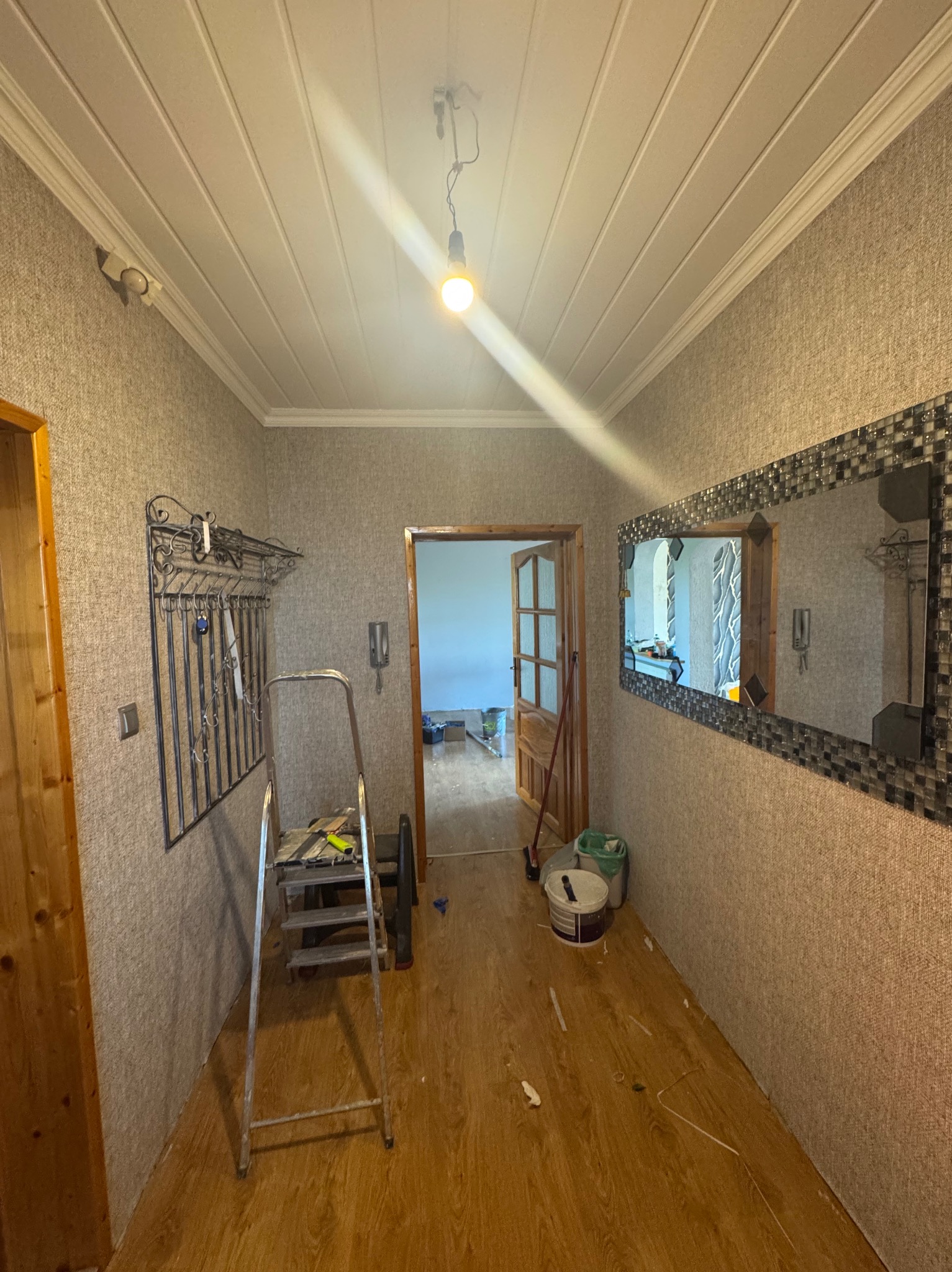 Korytarz w trakcie prac wykończeniowych: drabina, pędzle, wiadro z farbą. Dekoracyjne lustro i wieszak na ścianie. Widoczne niedociągnięcia na podłodze.