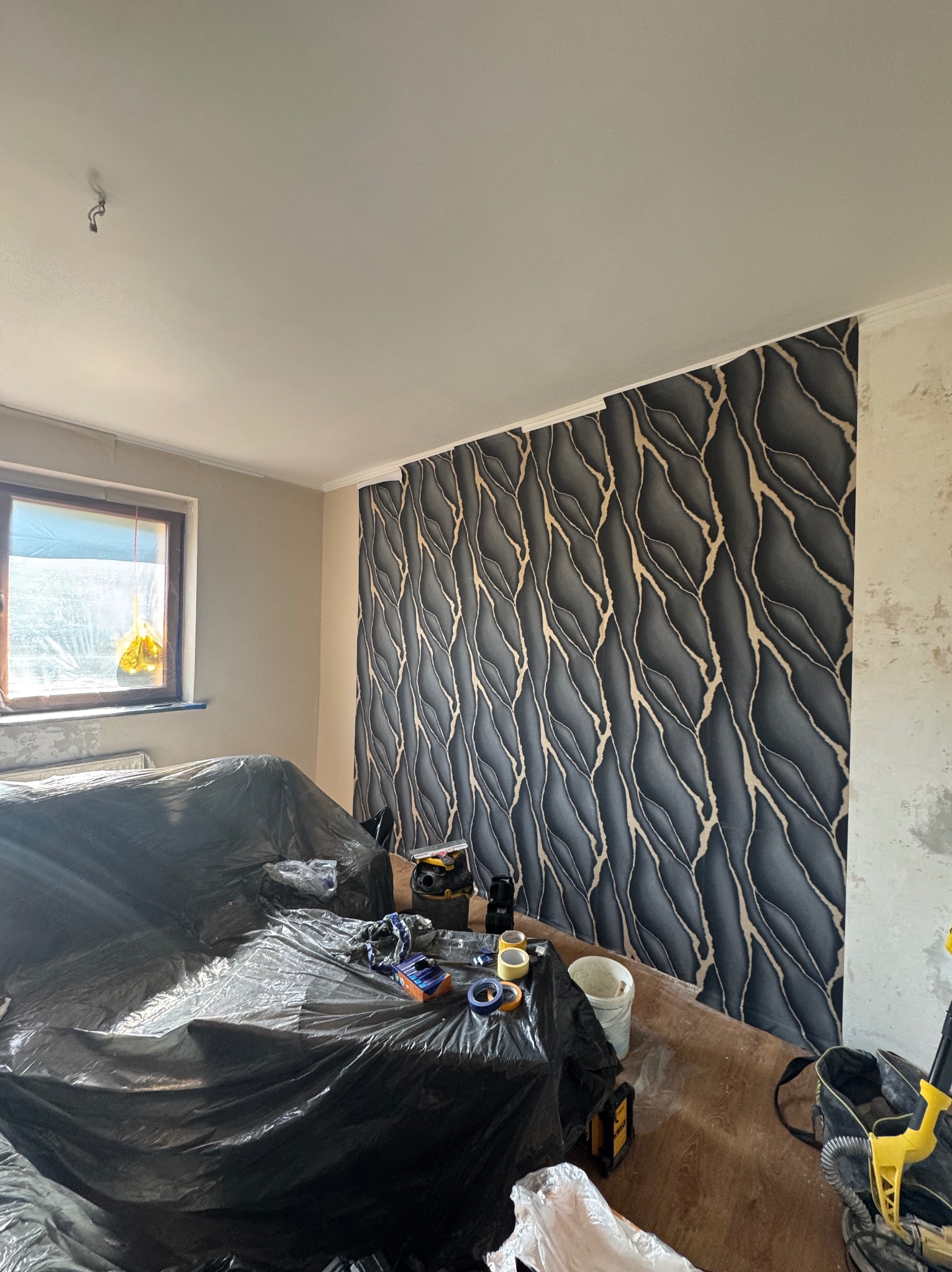 Ściana z tapetą w odcieniach szarości i złota, zabezpieczone meble folią, narzędzia malarskie widoczne na podłodze, fragment ściany przed tapetowaniem.