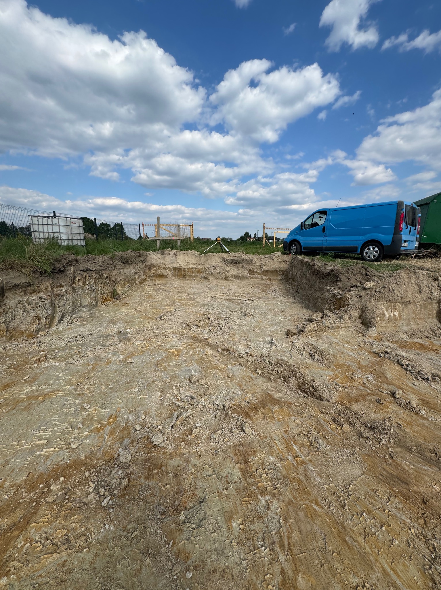 Wykonany wykop pod fundamenty domu jednorodzinnego, z boku zaparkowany niebieski samochód dostawczy. W tle widoczne niebo z chmurami.