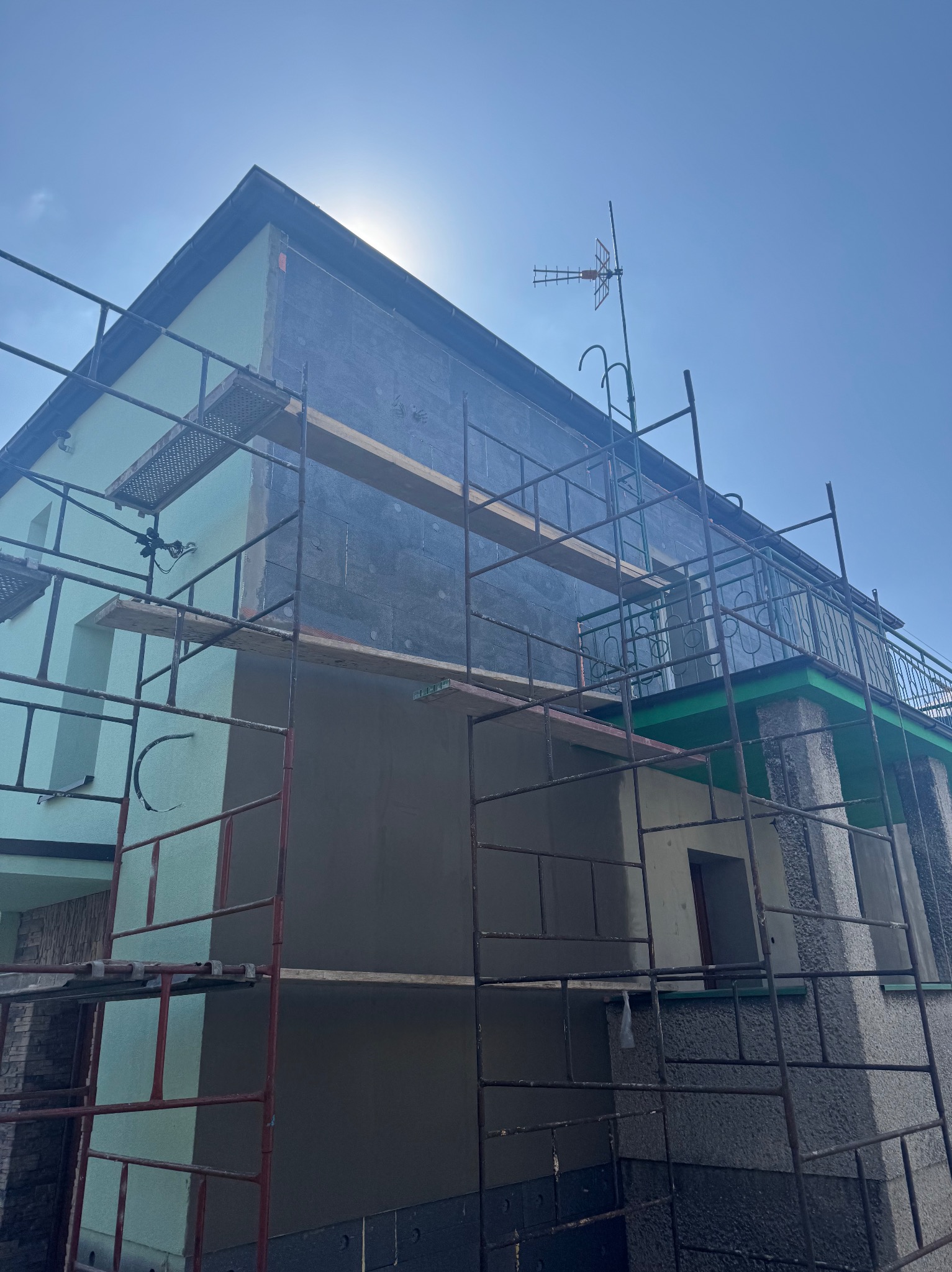 Dom w trakcie prac elewacyjnych: ocieplenie grafitowym styropianem, fragment otynkowany, rusztowania, antena na dachu, słoneczny dzień.