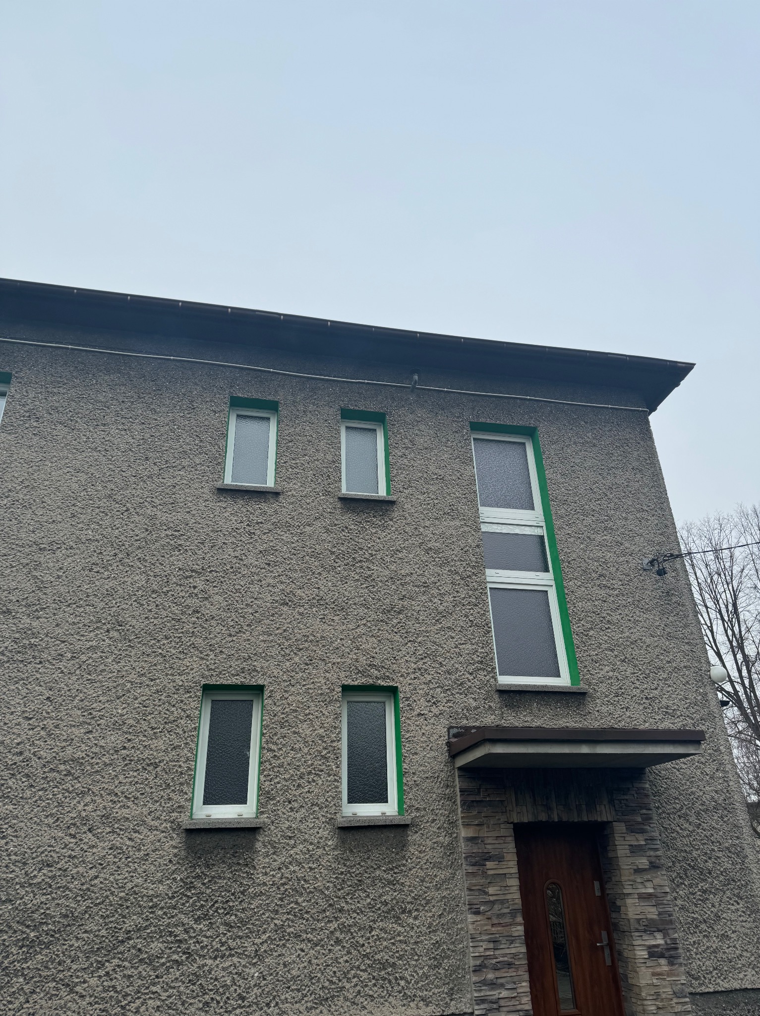 Szara elewacja budynku z oknami w jasnych, zielonych ramach i wejściem z kamienną okładziną. Chropowata faktura tynku, pochmurne niebo w tle. Ujęcie z dołu.