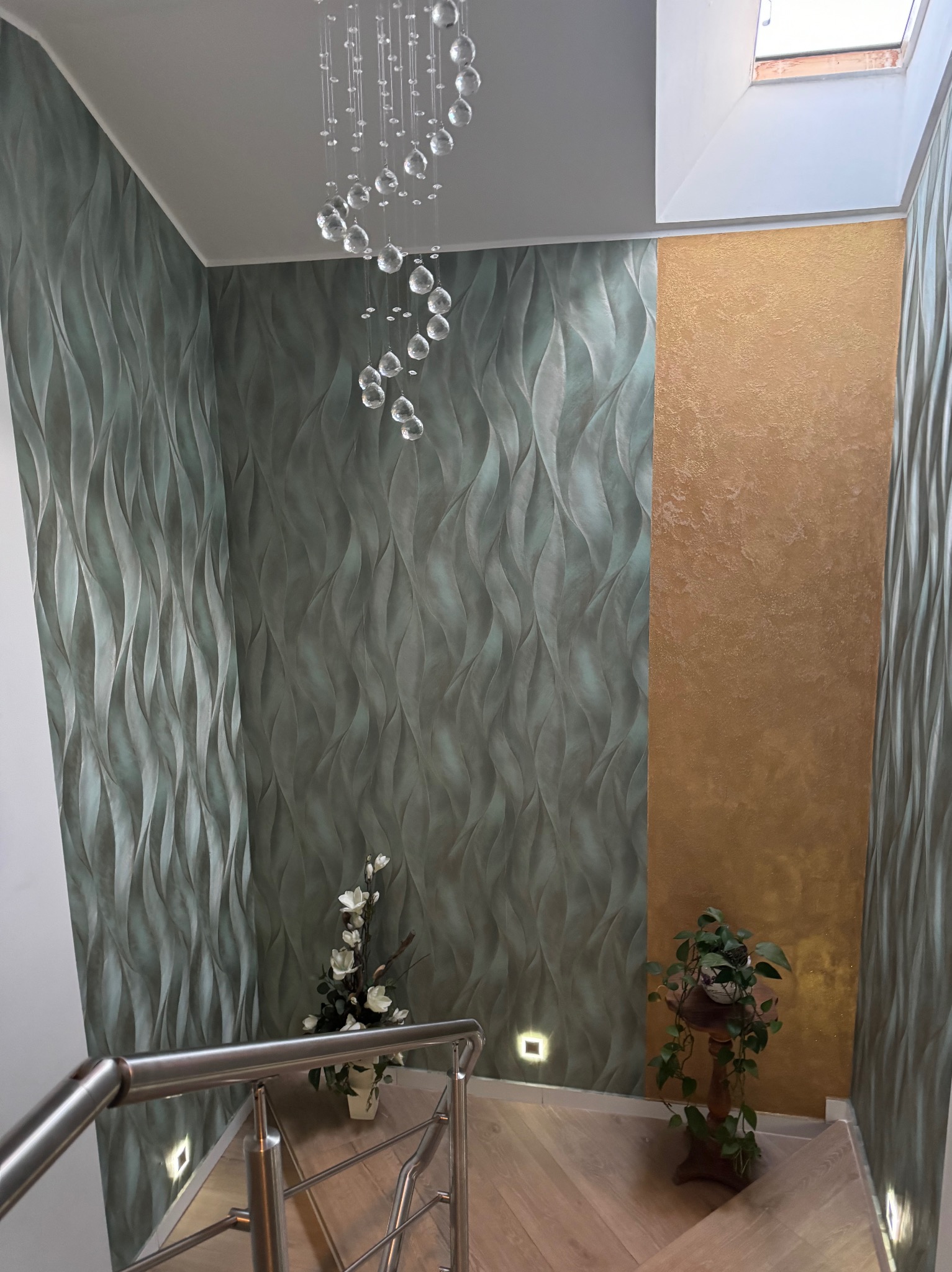 Klatka schodowa z dekoracyjną, trójwymiarową fakturą na ścianach, oświetleniem punktowym i nowoczesnym żyrandolem. Widoczny fragment schodów i rośliny doniczkowe.