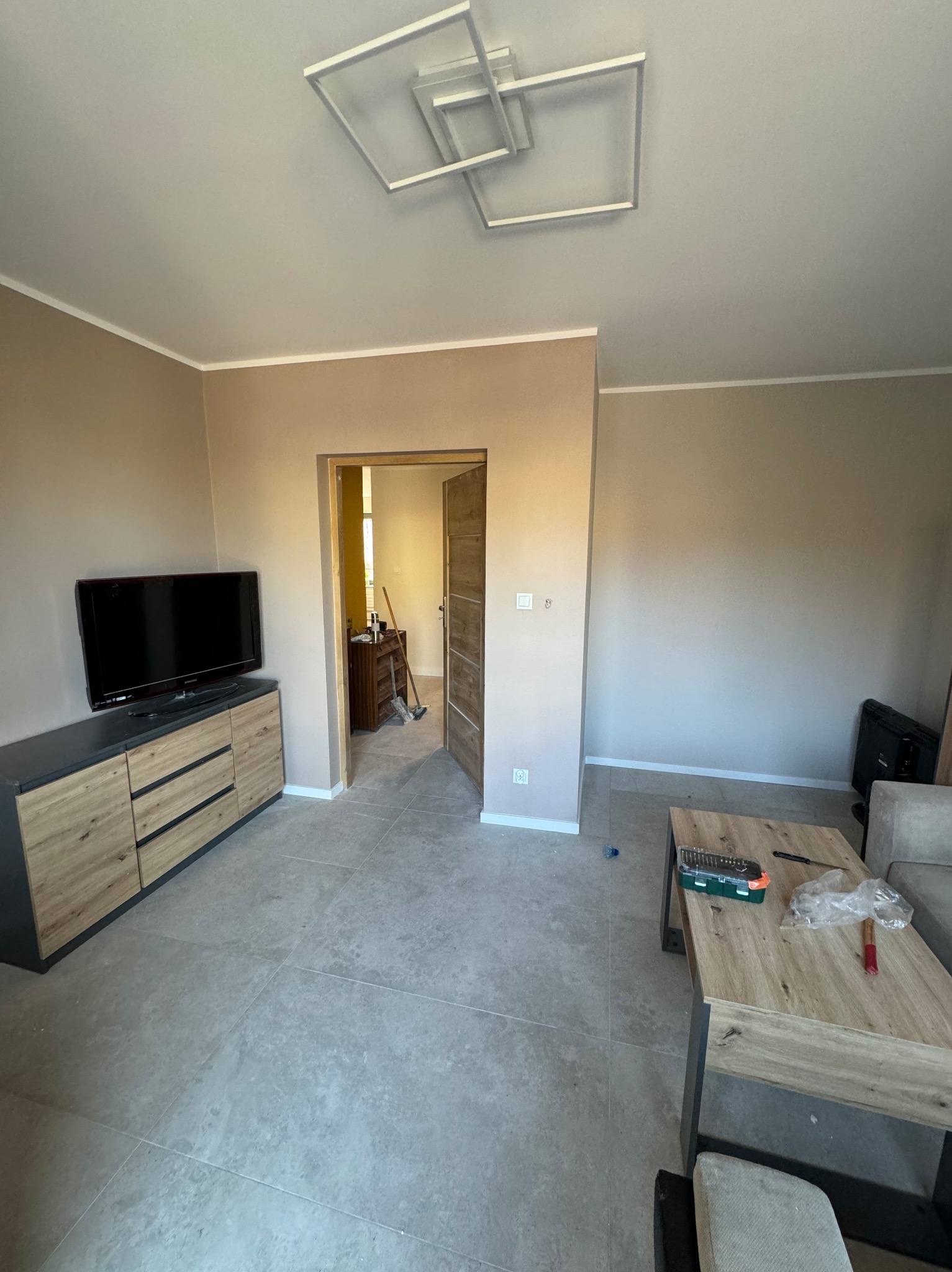 Beżowe ściany w pokoju po malowaniu, widoczny fragment mebli, podłogi i sufitu z nowoczesną lampą. W tle otwarte drzwi do innego pomieszczenia.