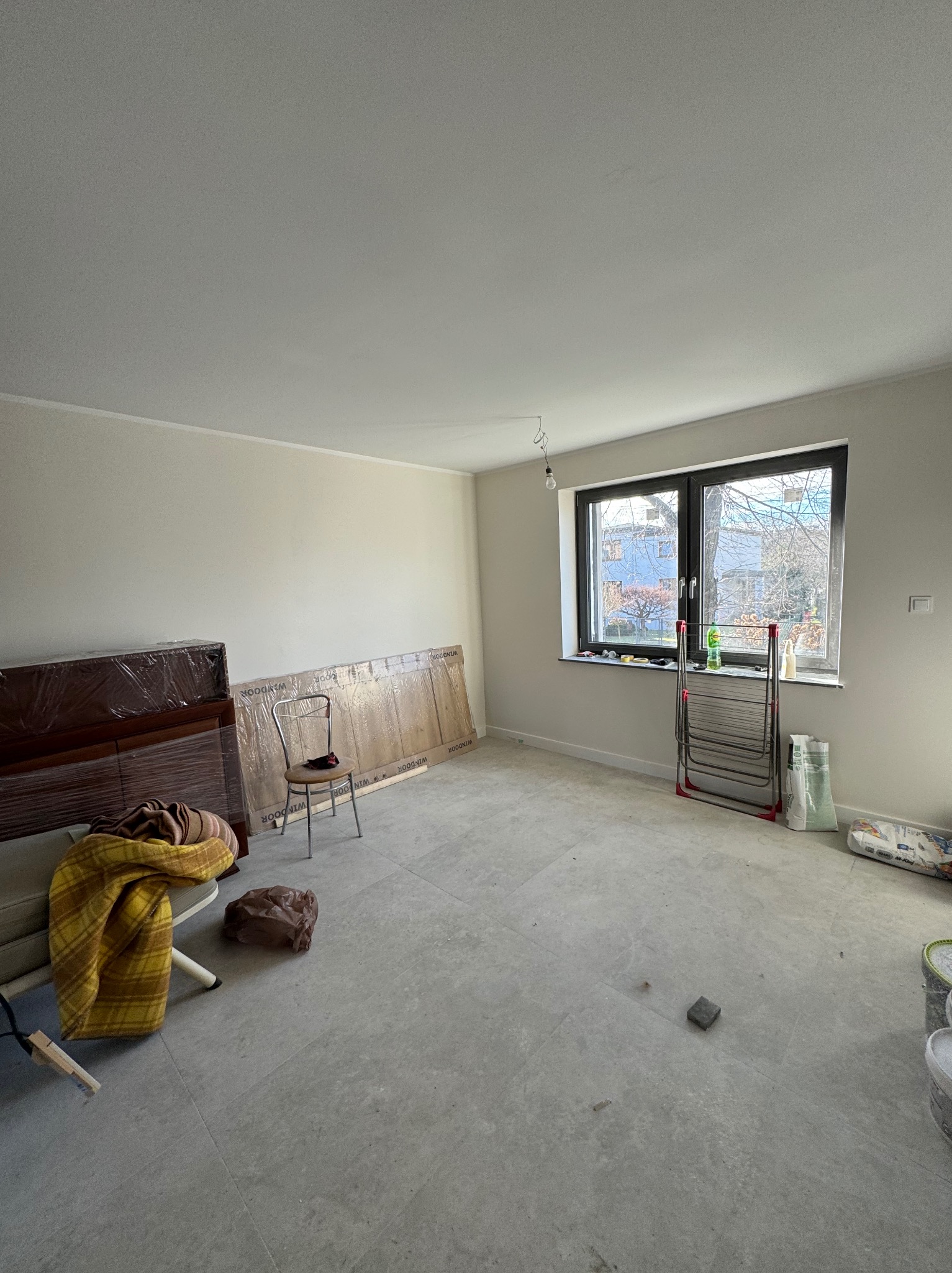 Jasne, puste wnętrze w trakcie remontu: szare płytki podłogowe, okno z widokiem na zewnątrz, meble zabezpieczone folią i drabina do suszenia prania.