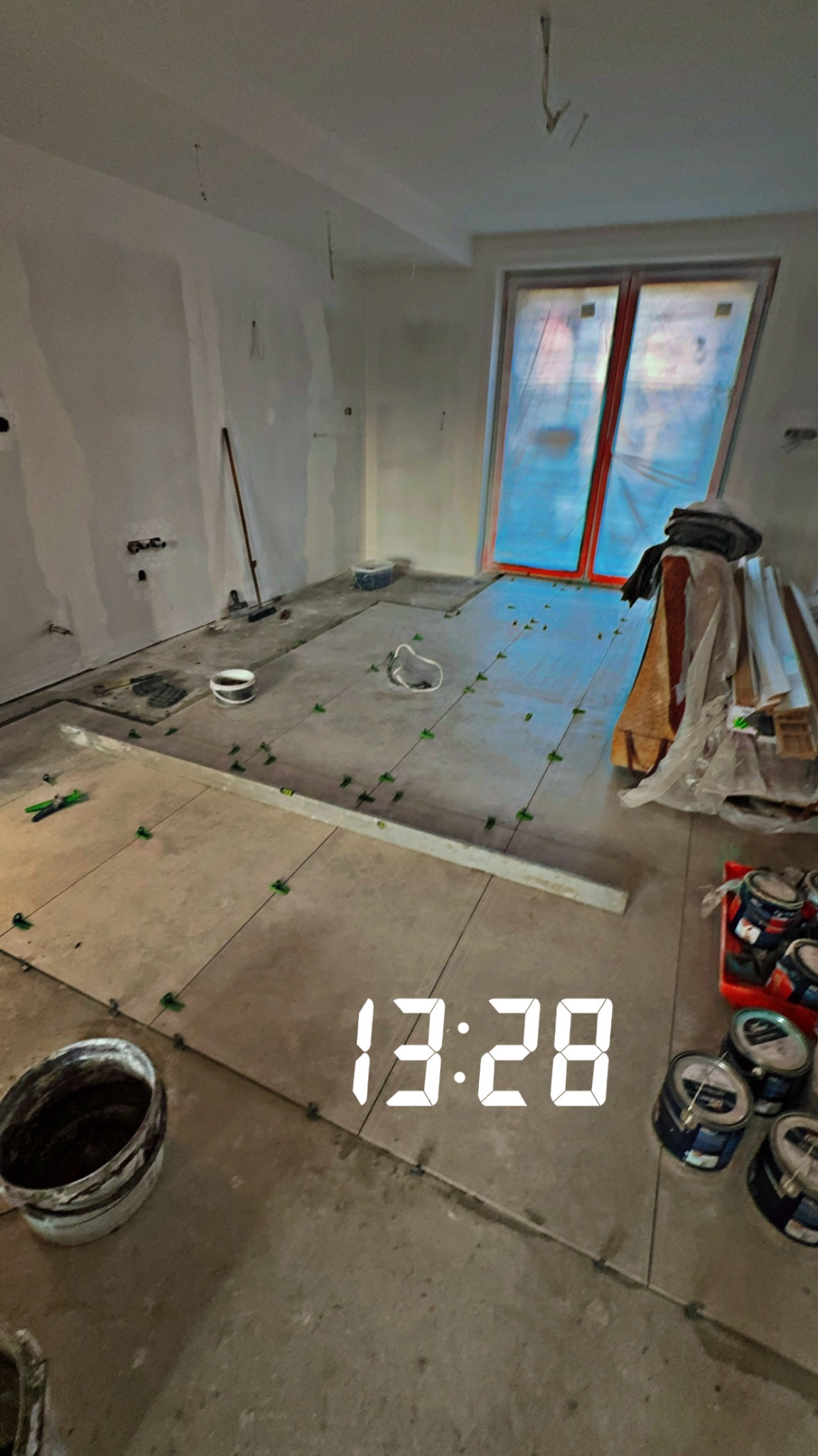 Układanie płytek podłogowych w jasnym wnętrzu, z widocznymi krzyżykami dystansowymi, wiadrem z zaprawą i puszkami farby. Okno zabezpieczone folią.