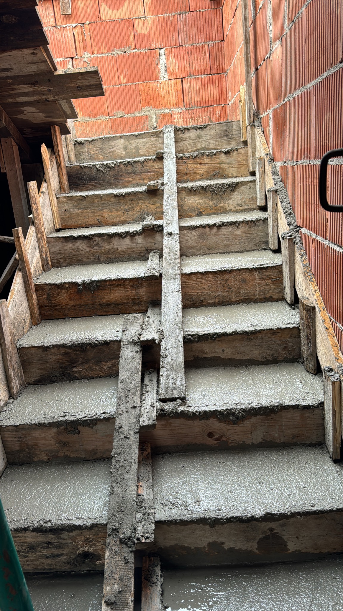 Betonowe schody w drewnianym szalunku, widok z dołu. Świeży beton wypełnia stopnie, ceglana ściana z boku. Etap budowy schodów wewnętrznych.