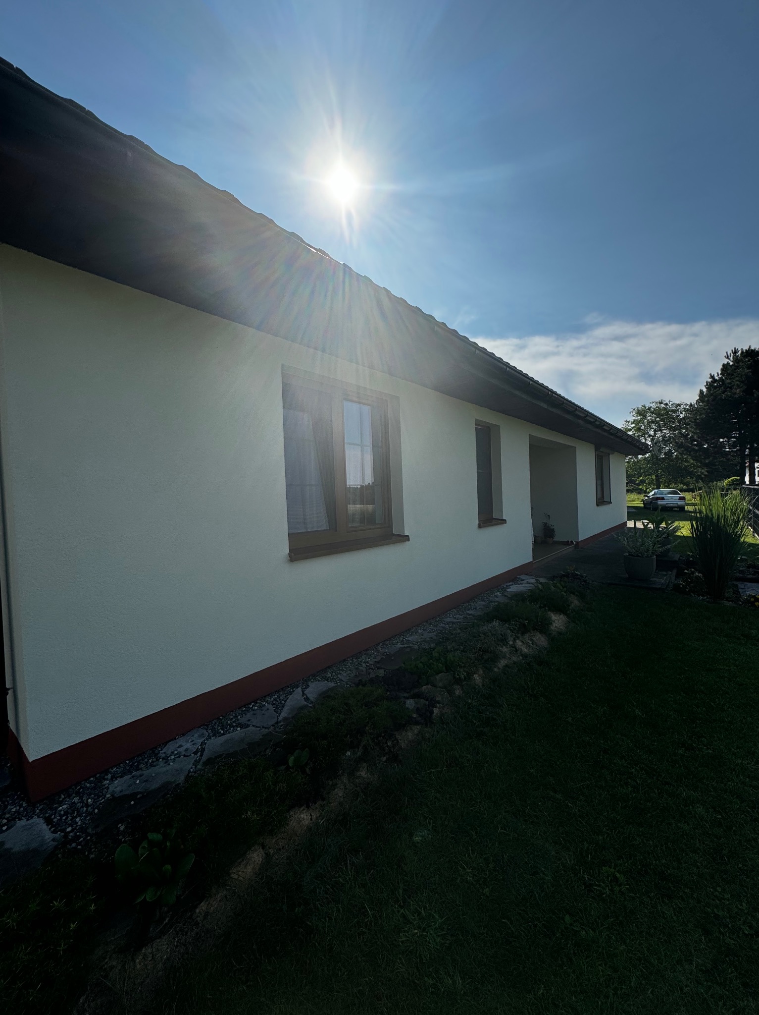 Boczny widok domu po malowaniu elewacji w słoneczny dzień. Biała fasada, brązowe okna i podmurówka. Zieleń trawy i roślinności. Błękitne niebo z promieniami słońca.