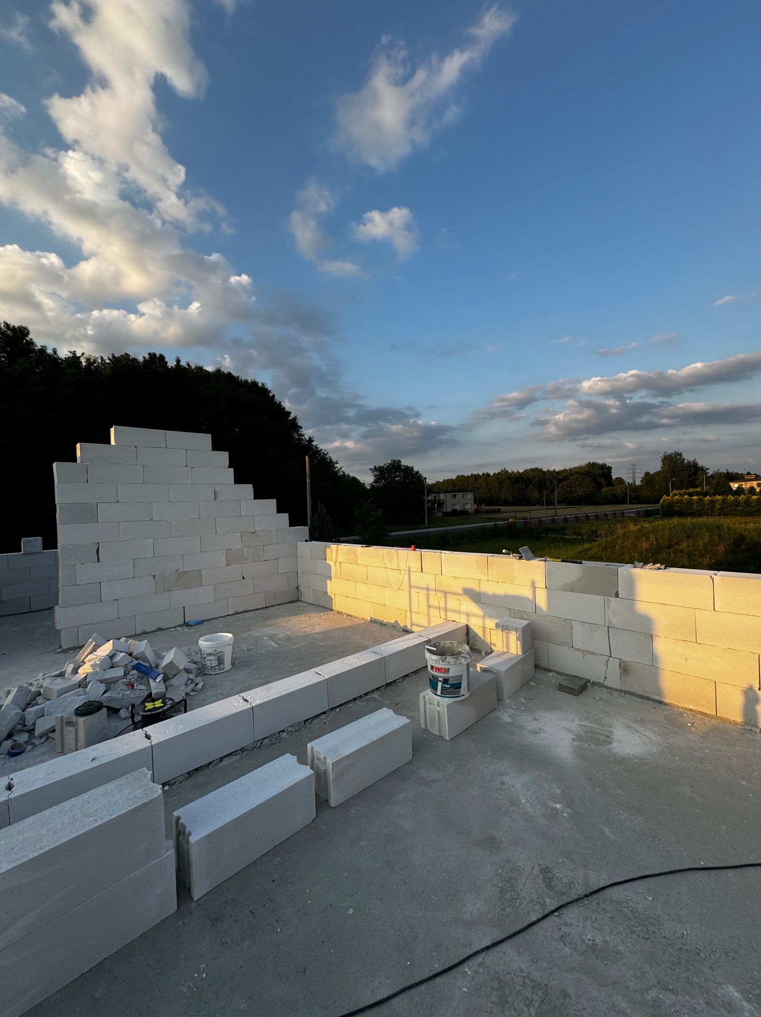 Wznoszenie ścian z białych bloczków betonu komórkowego na budowie domu jednorodzinnego, widok na las i błękitne niebo z chmurami o zachodzie słońca.