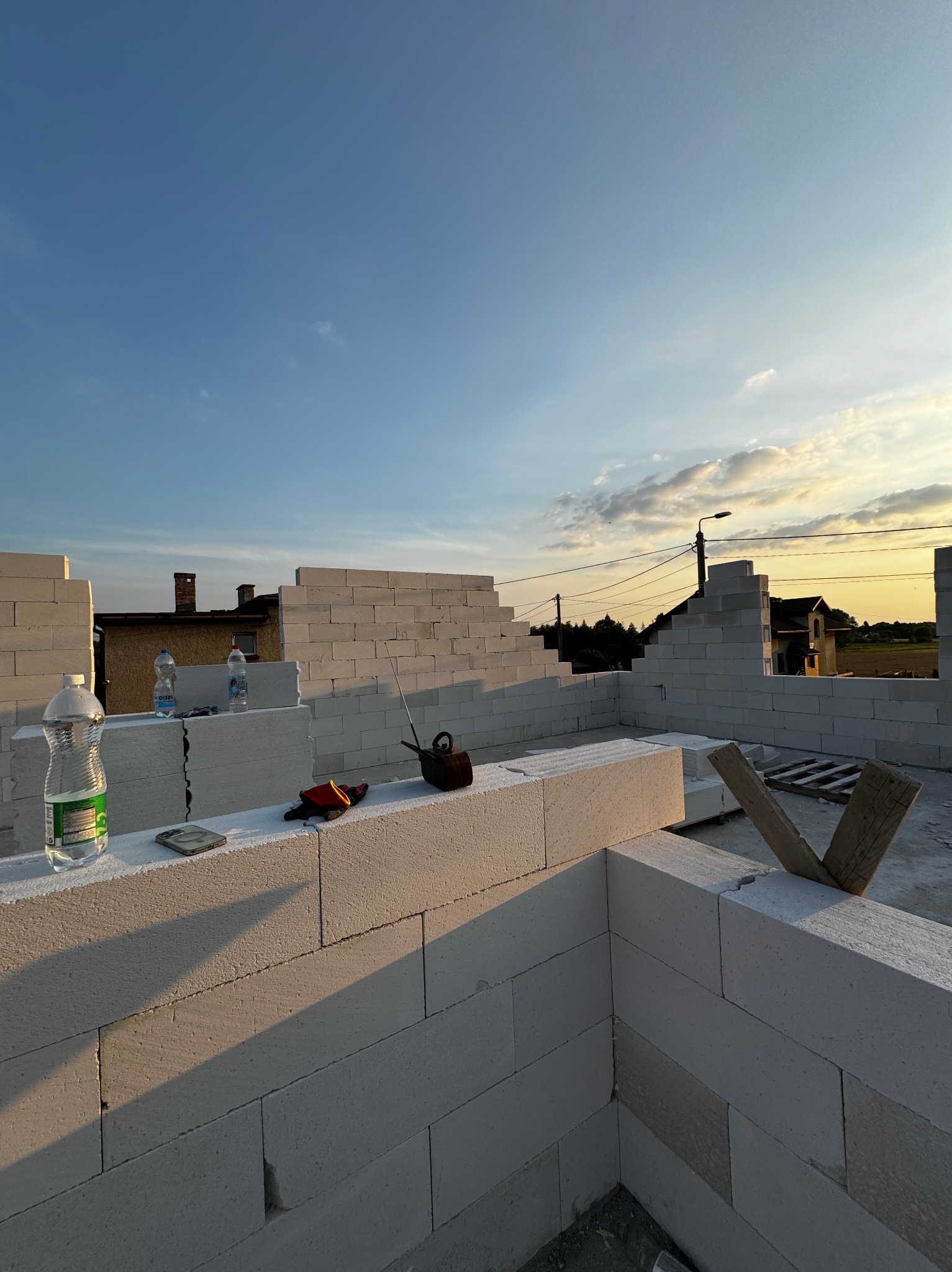 Wznoszone ściany domu z białych bloczków, widoczne narzędzia, butelki z wodą i elementy drewniane. W tle błękitne niebo i zabudowania.