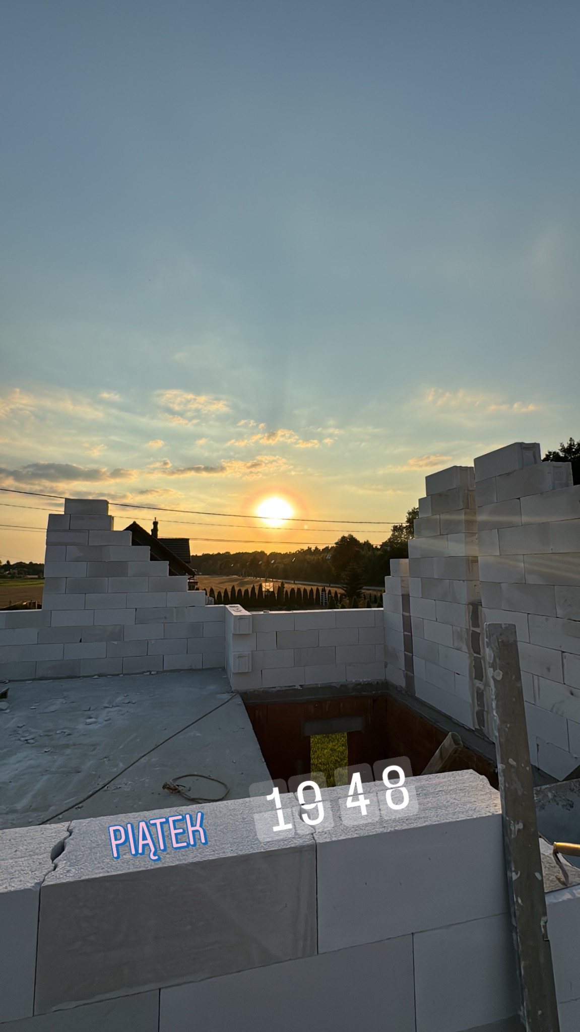 Wznoszone ściany z białych bloczków na tle zachodzącego słońca. Widoczny murarski poziom i fragment otworu okiennego. Na pierwszym planie napis 'Piątek'.