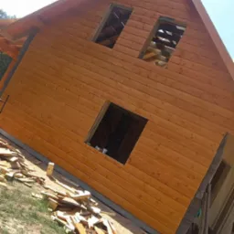 Drewniany dom w trakcie budowy, z widocznymi otworami okiennymi i stertą drewnianych odpadów na trawie, świeżo pomalowana elewacja, jasne oświetlenie.