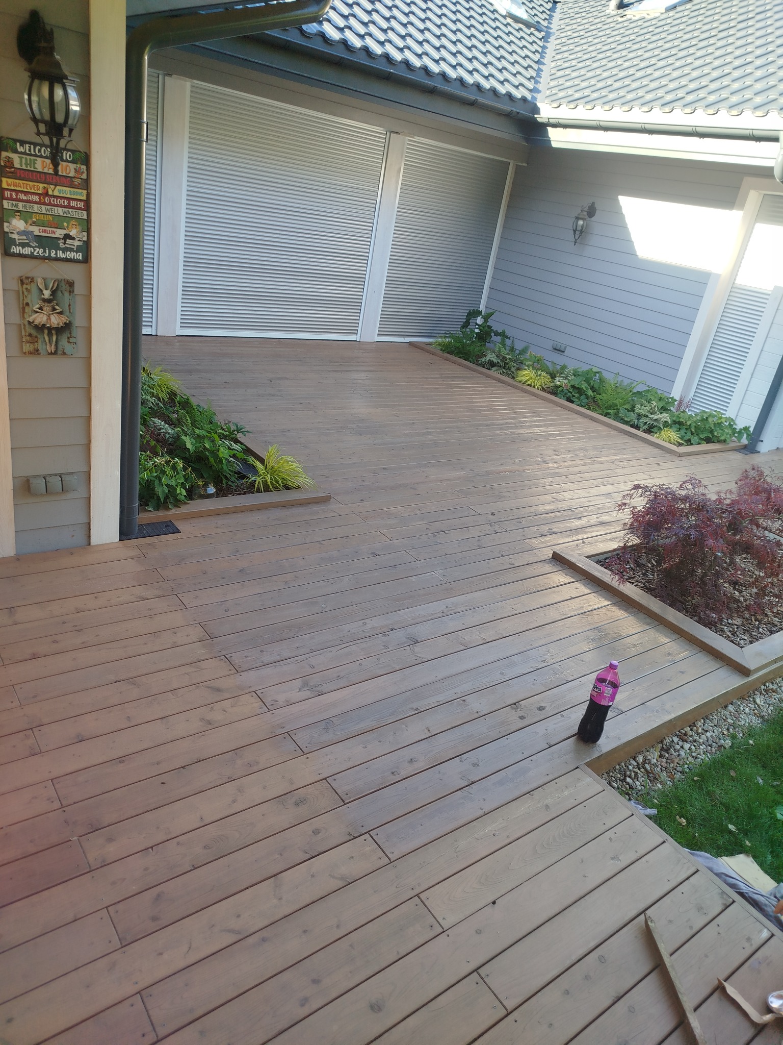 Odnowiony drewniany taras przylegający do domu z szarą elewacją i roletami zewnętrznymi. Widoczne rośliny ozdobne i dekoracje.