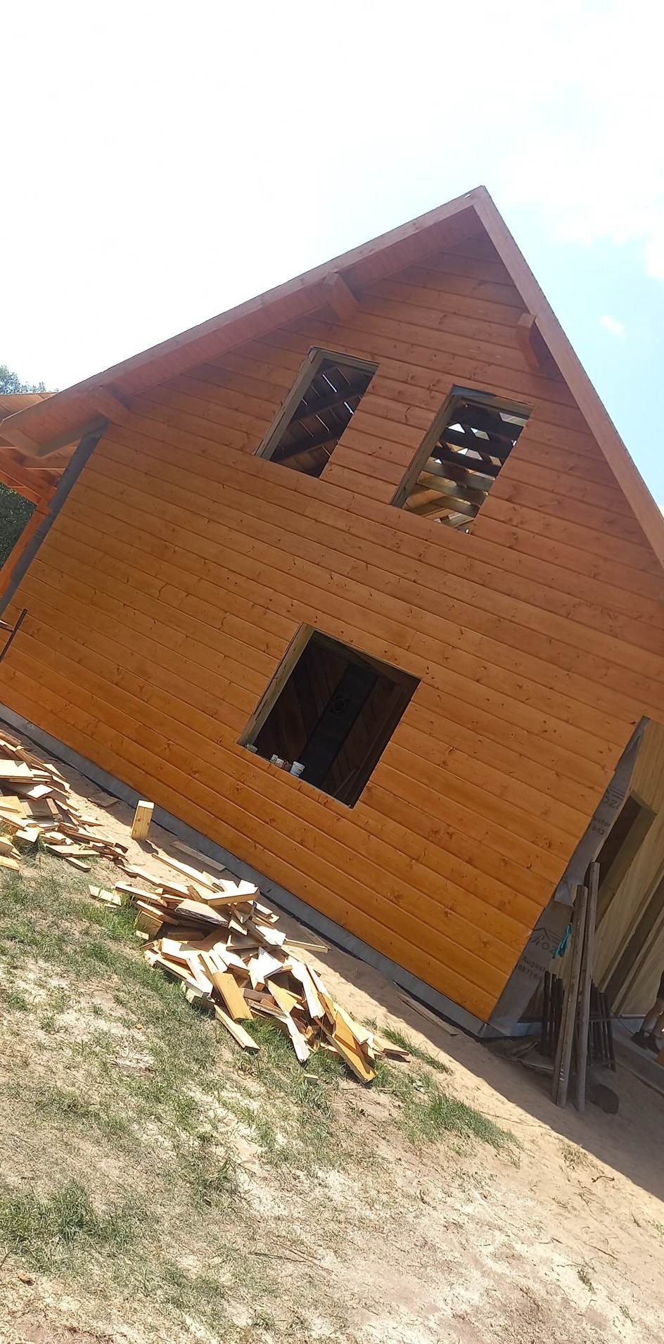 Drewniany dom w trakcie budowy, z widocznymi otworami okiennymi i stertą drewnianych odpadów na trawie, świeżo pomalowana elewacja, jasne oświetlenie.