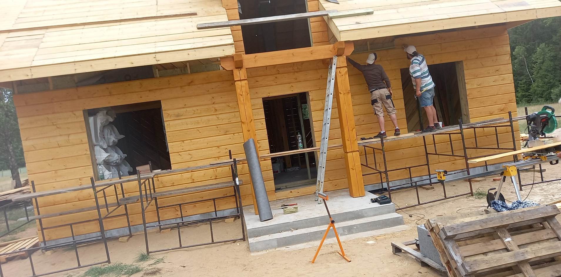 Budowa domu z drewnianą elewacją w trakcie realizacji. Dwóch pracowników na rusztowaniu montuje elementy. Widoczne narzędzia i materiały budowlane.