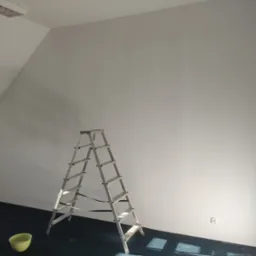 Wnętrze po malowaniu: białe ściany pod skosem, aluminiowa drabina, miska z farbą na ciemnej wykładzinie. Widoczny kontakt elektryczny.