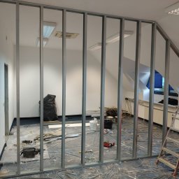 Szymon Bulczak - Montaż metalowego szkieletu ścianki działowej w jasnym pokoju na poddaszu, widoczne narzędzia i materiały budowlane, drabina. Folia ochronna na podłodze.