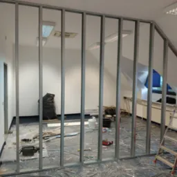 Montaż metalowego szkieletu ścianki działowej w jasnym pokoju na poddaszu, widoczne narzędzia i materiały budowlane, drabina. Folia ochronna na podłodze.