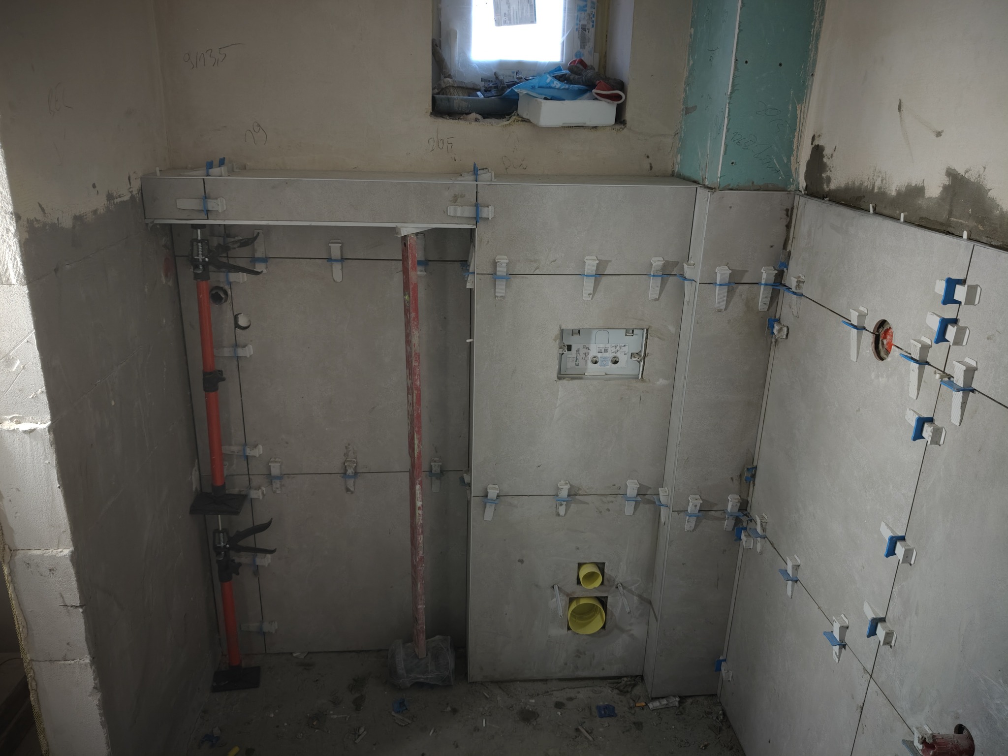 Szare płytki ścienne, w trakcie montażu, z użyciem niebieskich krzyżyków dystansowych i poziomicy laserowej. Widoczne otwory na instalacje. Surowe wnętrze łazienki w trakcie wykończenia.