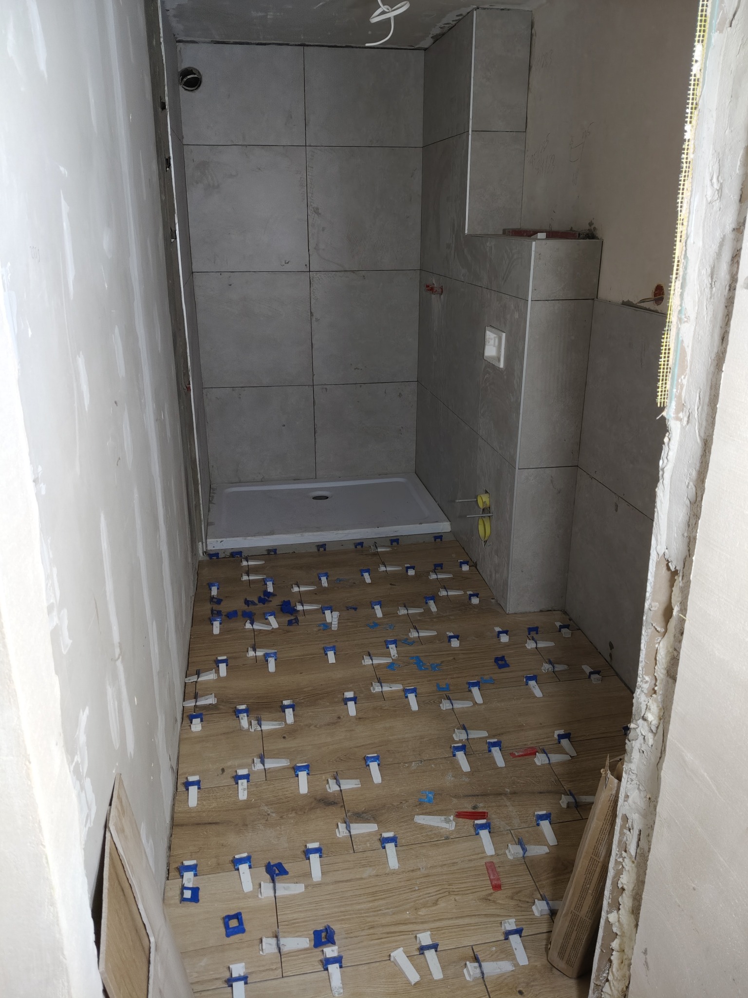 Łazienka w trakcie wykańczania: ułożone płytki imitujące drewno na podłodze, szare płytki na ścianach, widoczny brodzik. Użyto klipsów poziomujących.