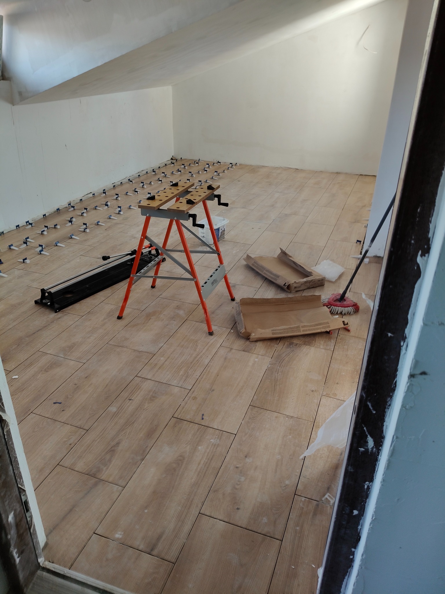 Układanie płytek podłogowych w skos na poddaszu. Widoczny stół roboczy, poziomica i system poziomowania płytek. Płytki imitują drewno, ułożone w jodełkę.