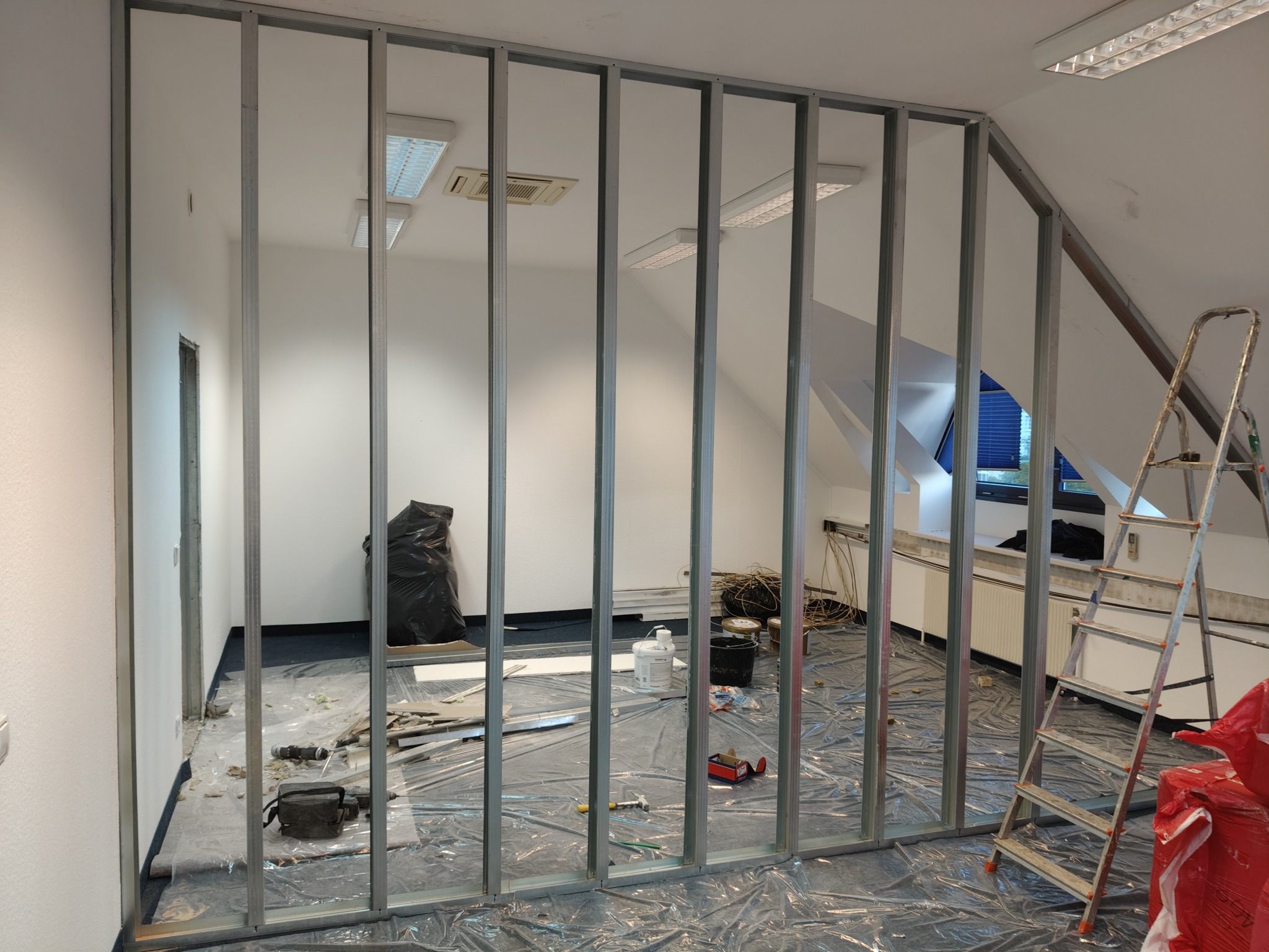 Montaż metalowego szkieletu ścianki działowej w jasnym pokoju na poddaszu, widoczne narzędzia i materiały budowlane, drabina. Folia ochronna na podłodze.